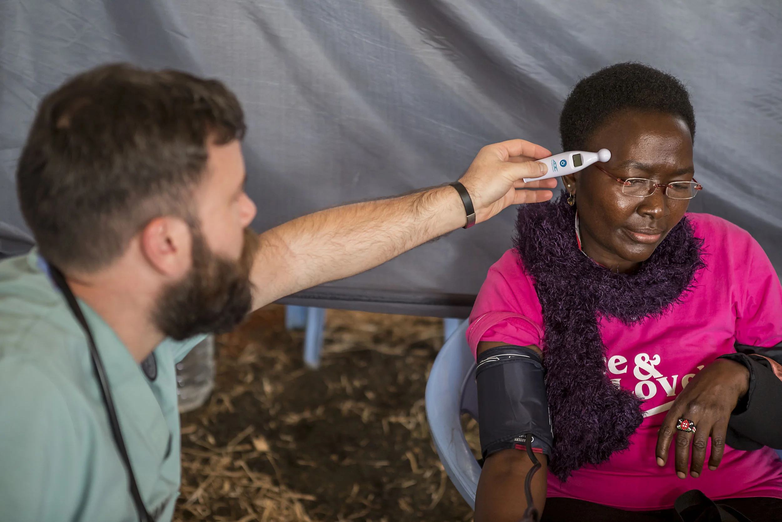 Man using a digital thermometer to take the temperature of a seated woman wearing a blood pressure cuff and pink shirt.