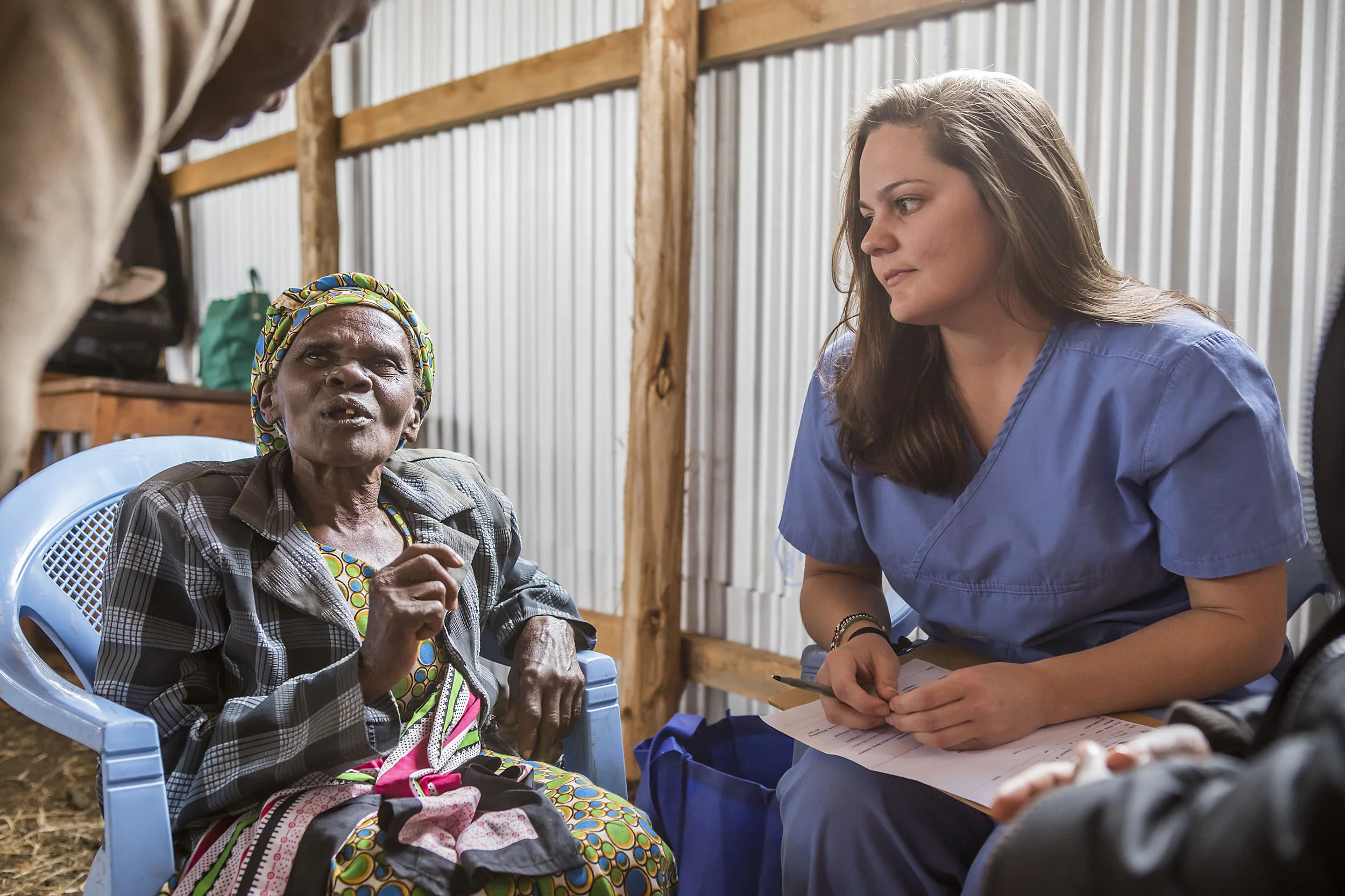 Healthcare worker in blue scrubs attentively listening to an elderly woman in colorful clothing inside a wooden and metal-paneled room.