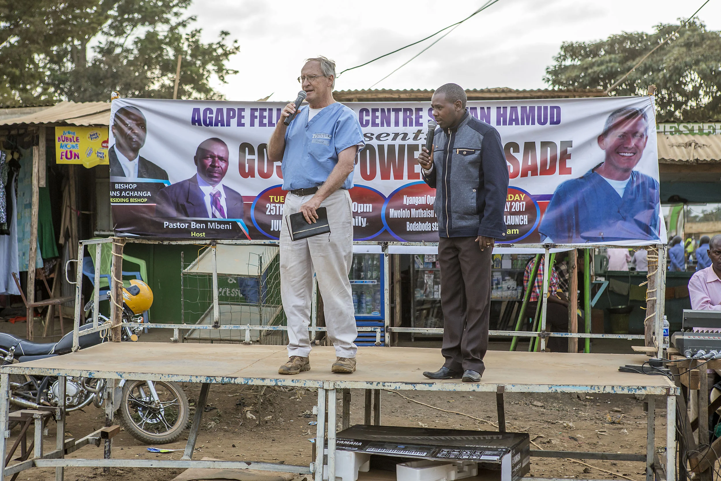 Two men holding microphones speaking on an outdoor stage with a banner promoting a gospel crusade event in the background.