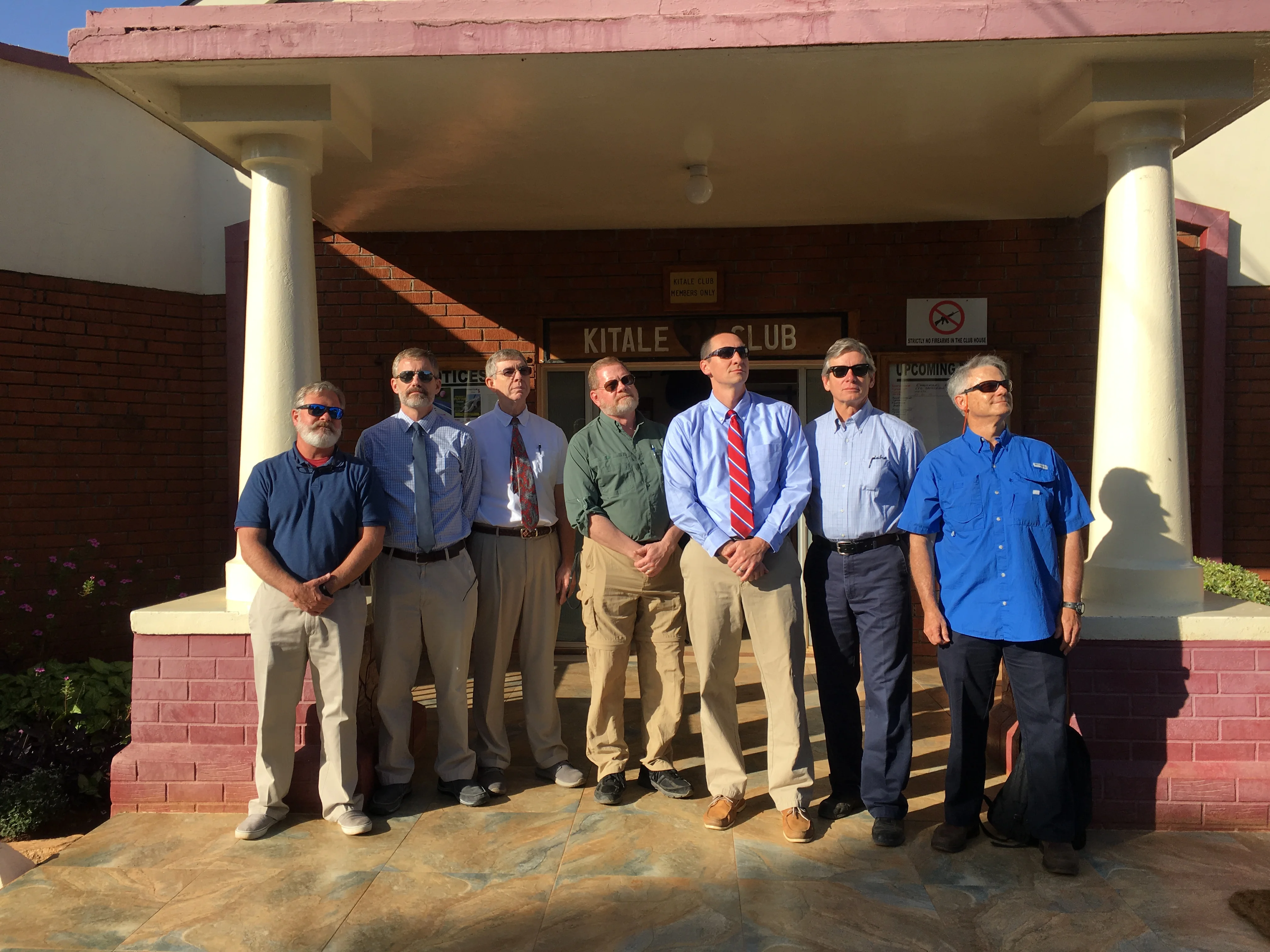 Seven men wearing sunglasses standing in a row outside a building with columns under a sign that reads 'Kitale Club'.