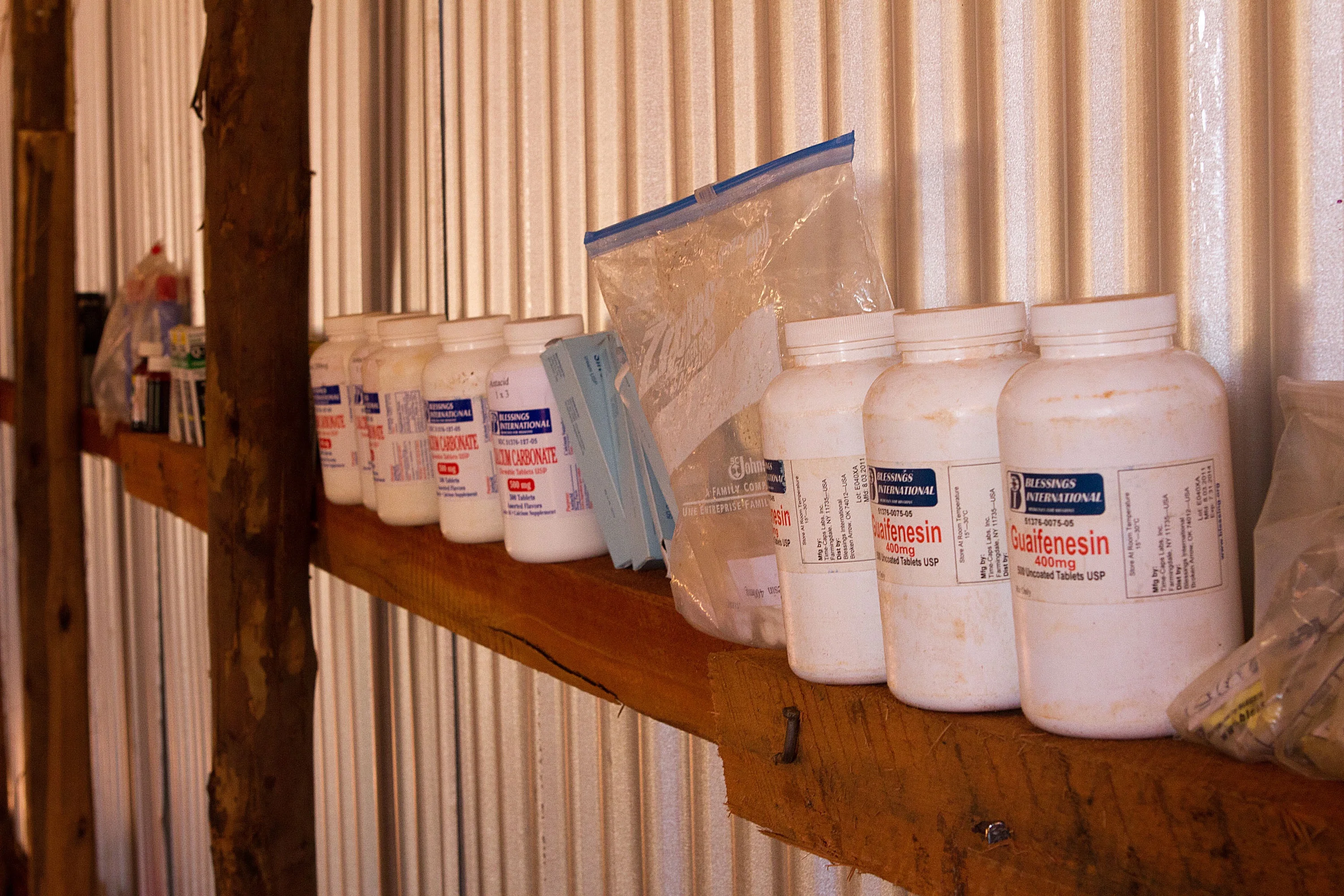 Row of white medication bottles labeled Guaifenesin and Calcium Carbonate on a wooden shelf against a corrugated metal wall.