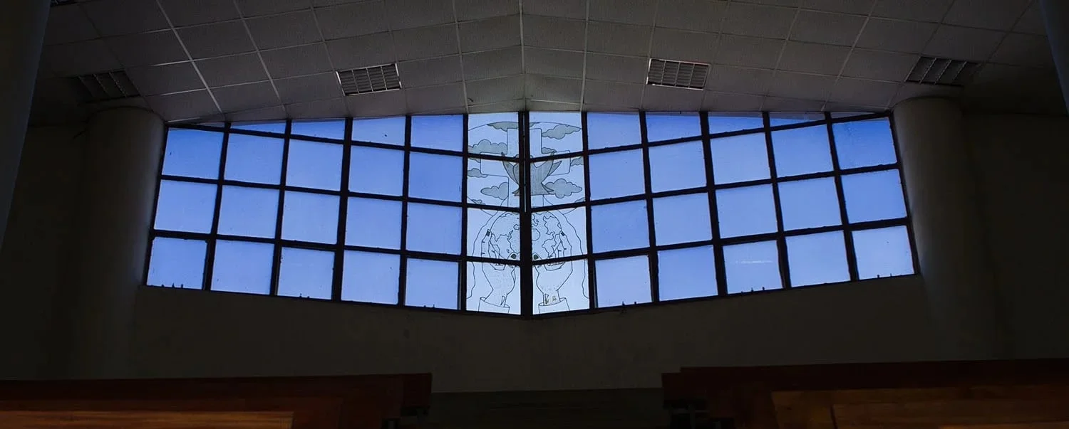 Large window with blue panes featuring an outline drawing of two hands holding the Earth with a tree and clouds above in a building interior.