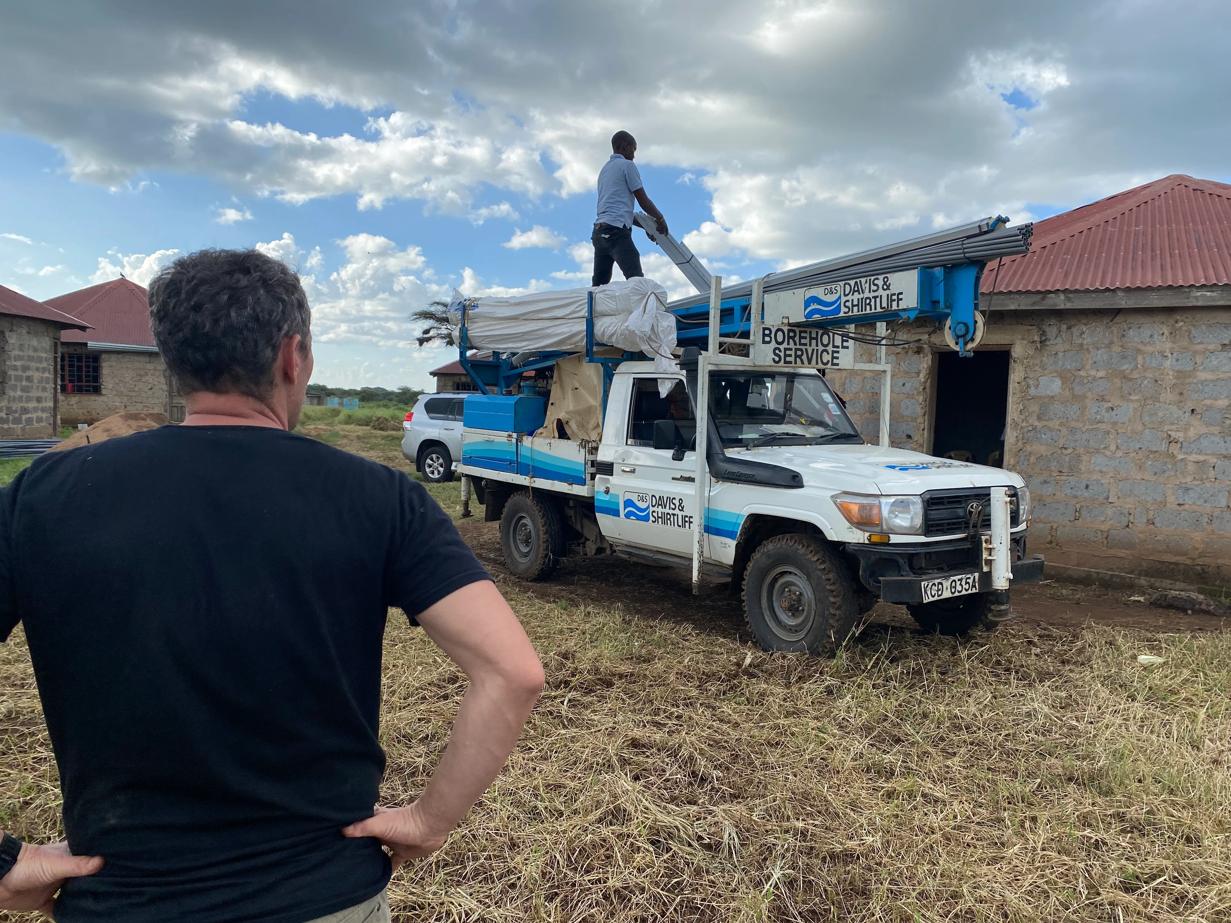 Man in black shirt observing another man standing on a white and blue borehole service truck with equipment, next to stone buildings with red roofs under a partly cloudy sky.