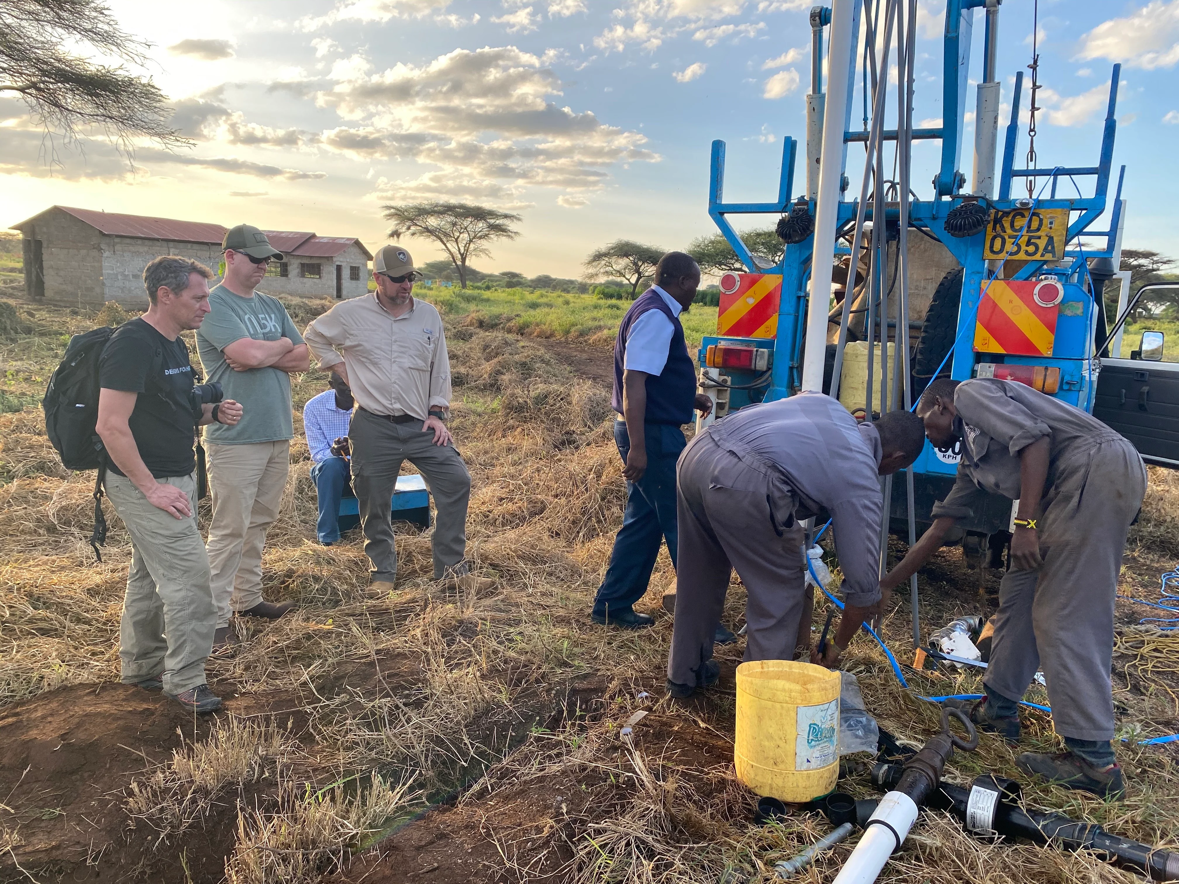 Group of men working with blue drilling equipment in a grassy field near small buildings at sunset.