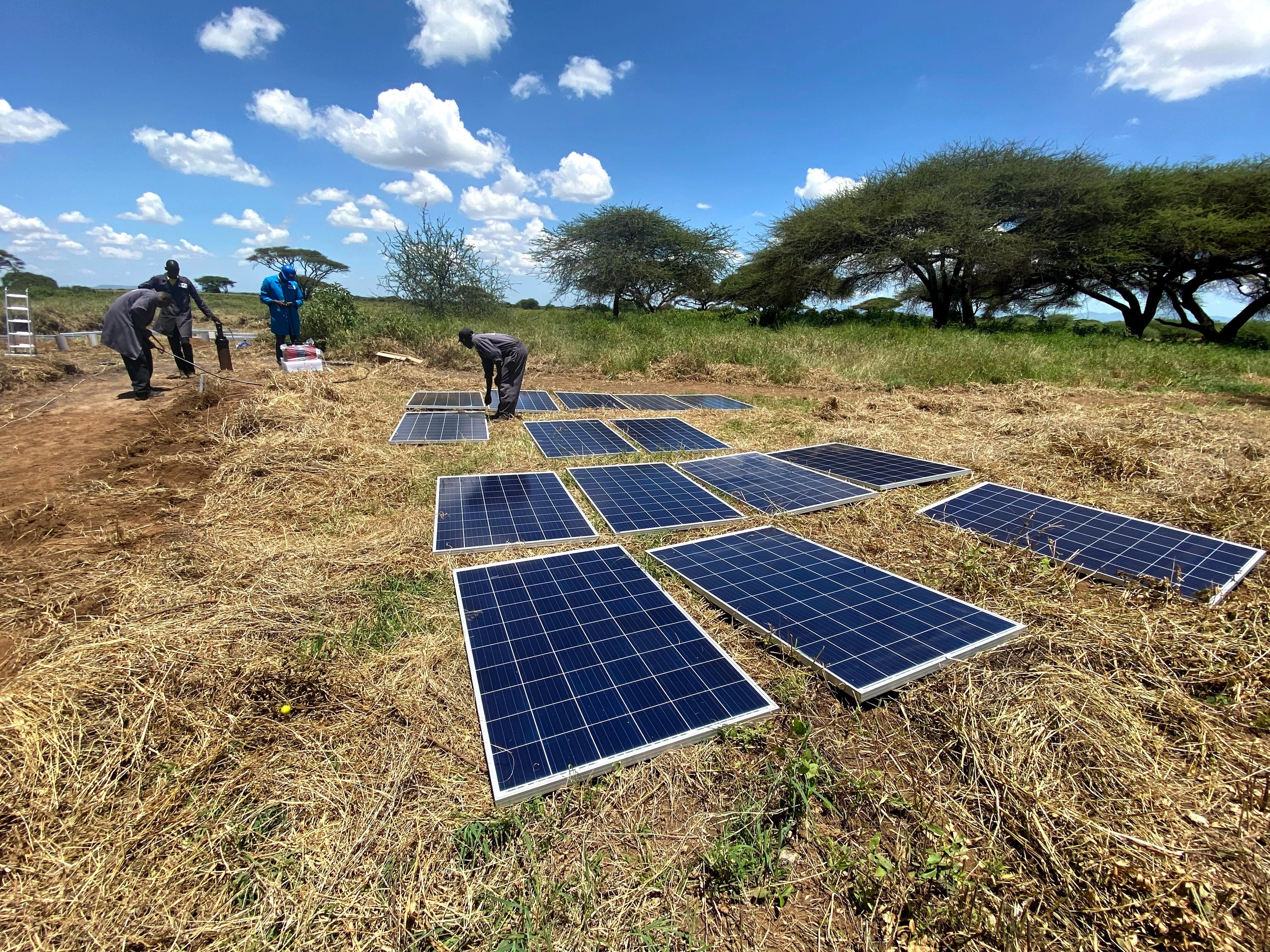 Workers installing solar panels on dry grassy land under a blue sky with scattered clouds.