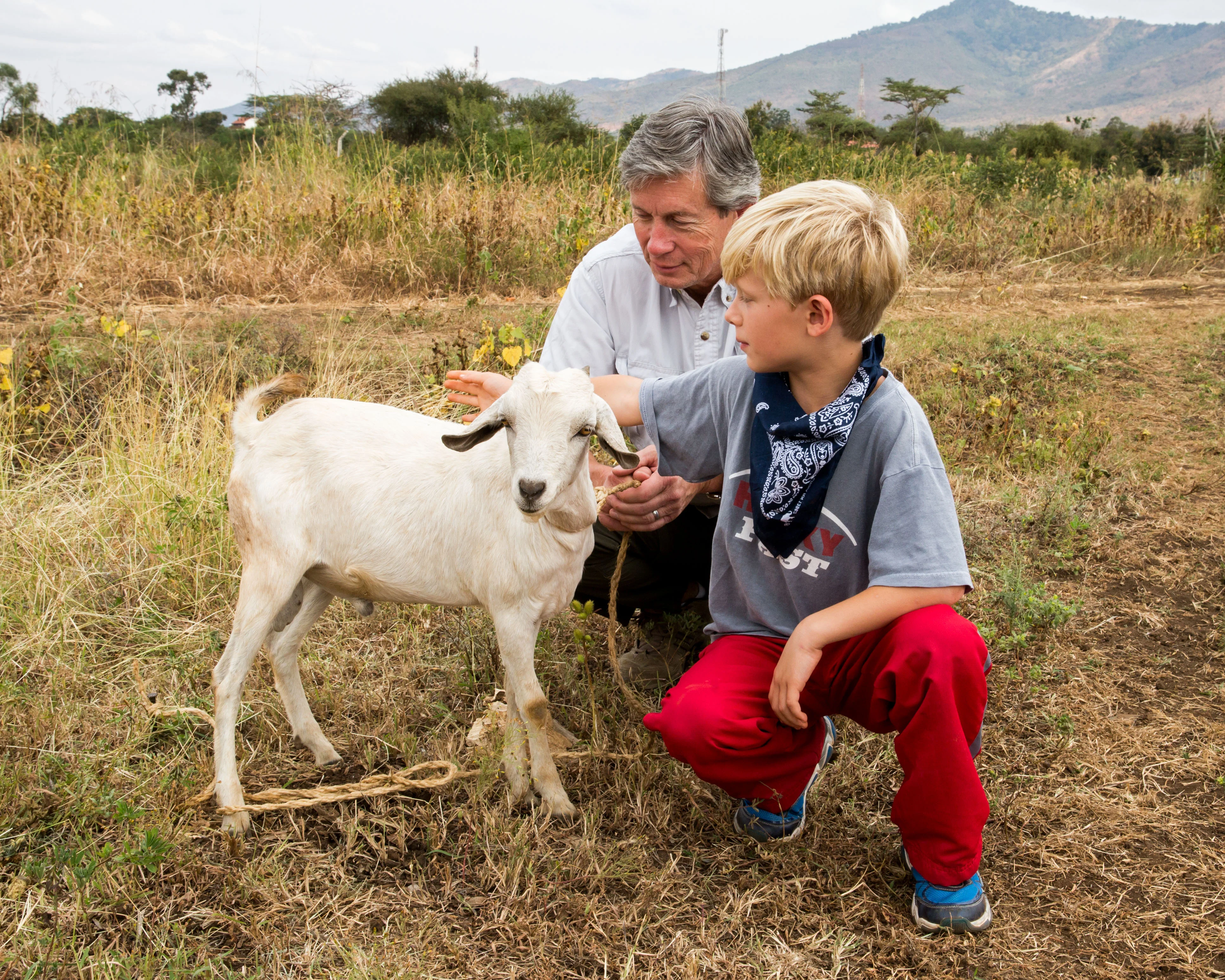 Young boy petting a white goat while an older man holds the goat's rope in a rural field with mountains in the background.