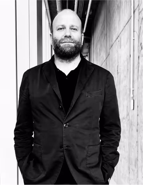 Martin Jasper de camisa negra y saco negro parado con las manos en los bolsillos en un pasillo de concreto