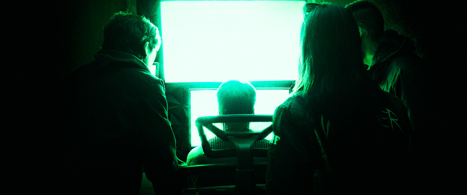 A moody dark room, with four people huddled around the bright green glow of computer screens