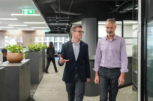 Two men in business attire walk and talk in a modern office hallway, with desks, plants, and another person visible in the background.