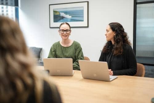 Three women sit at a conference table with laptops; one woman in glasses is speaking while the others listen. A framed photo hangs on the wall behind them.
