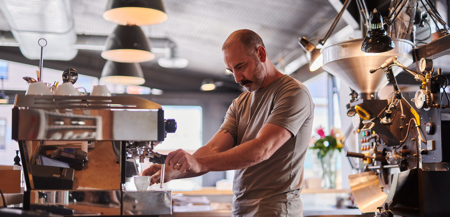 A man operates an espresso machine in a modern coffee shop, preparing a drink with various coffee equipment visible in the background.