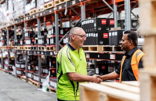 Two men wearing safety vests shake hands in a warehouse aisle lined with shelves of stacked boxes and pallets.