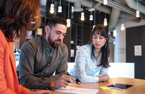 Three people sit at a table in a modern office, discussing documents and notes, with a tablet and sticky notes on the desk.