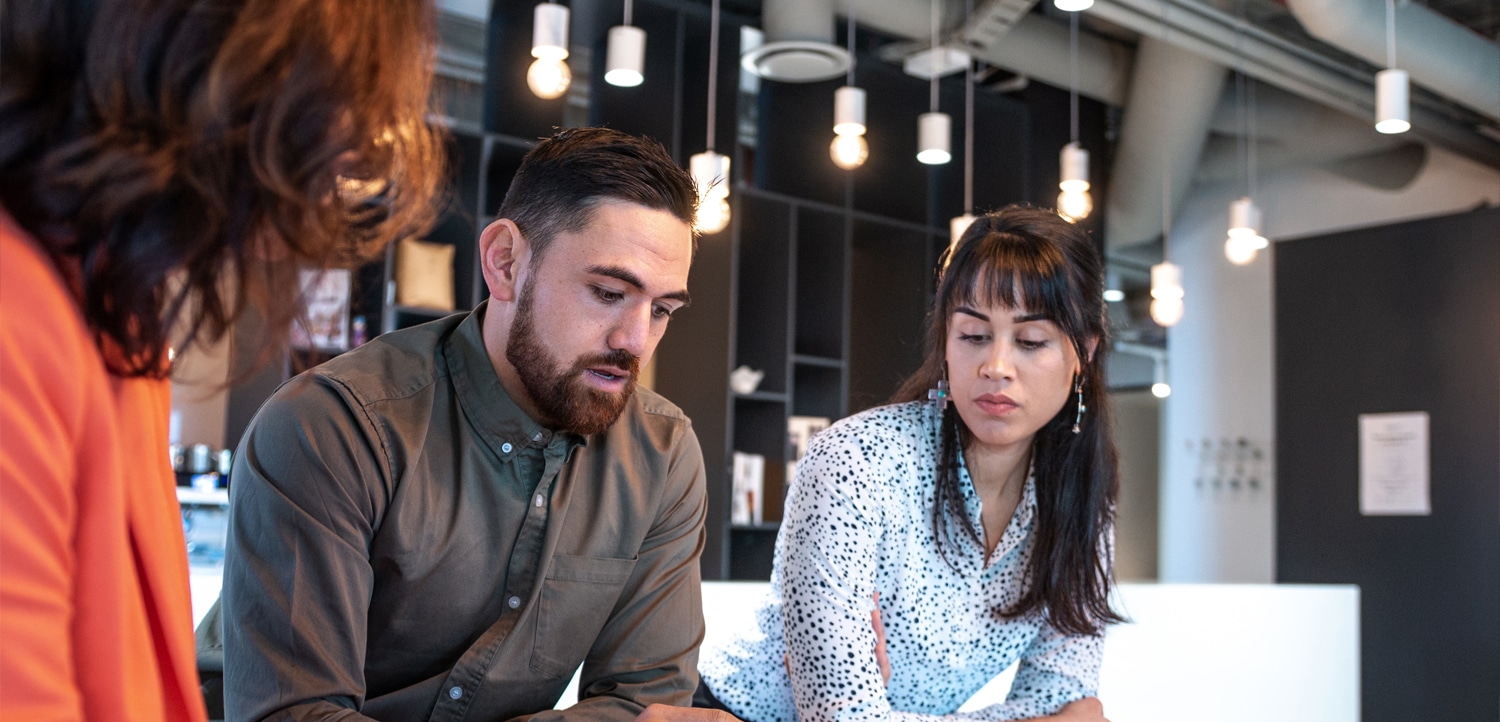 Three people sit at a table in a modern office, discussing documents and notes, with a tablet and sticky notes on the desk.