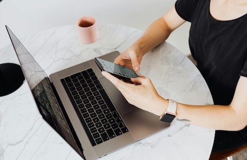Person using a smartphone while sitting at a round marble table with an open laptop and a pink mug nearby.