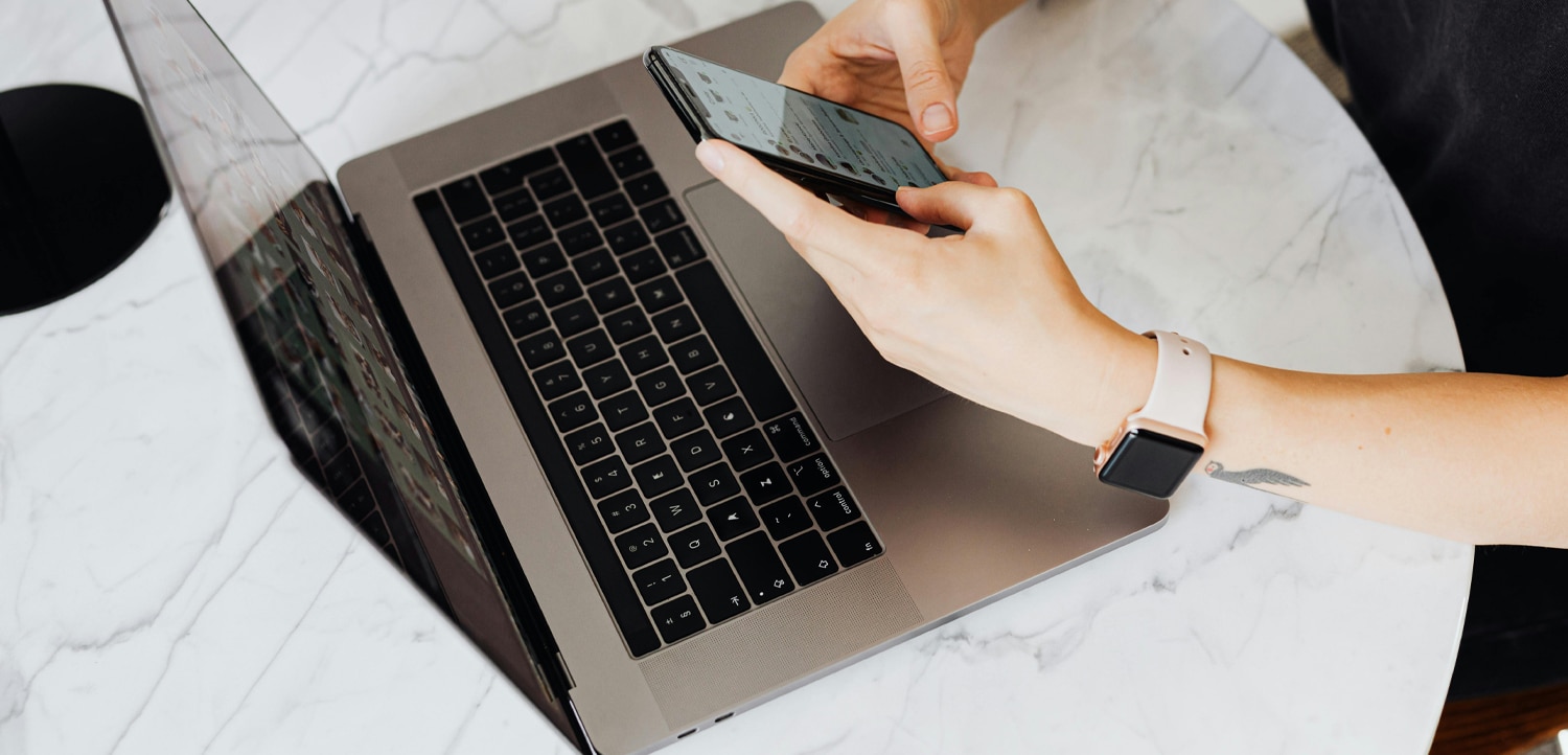 Person using a smartphone while sitting at a round marble table with an open laptop and a pink mug nearby.