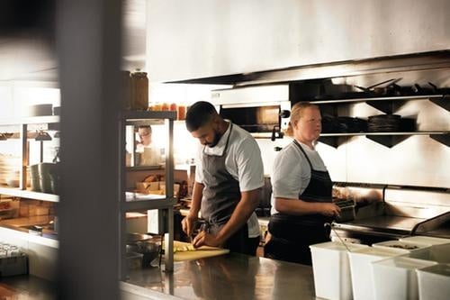 Two chefs work in a commercial kitchen; one chops vegetables while the other stands nearby, both wearing aprons and surrounded by kitchen equipment and ingredients.
