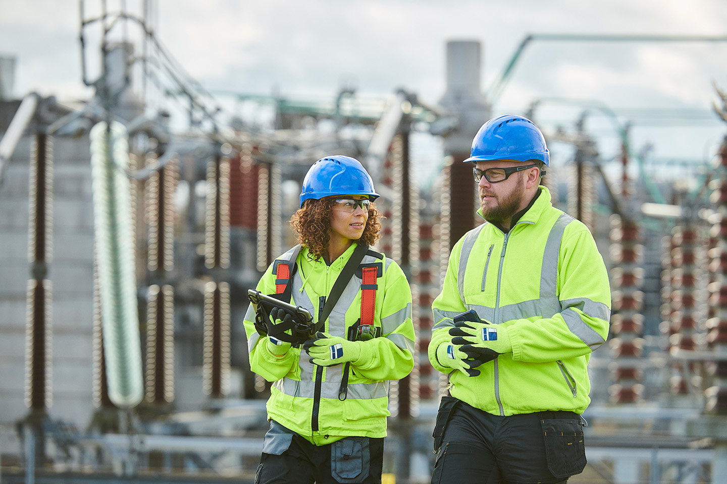 Two utility workers wearing blue helmets and neon yellow safety jackets inspecting equipment at an electrical substation.