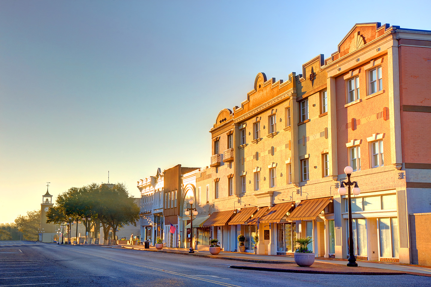 Sunlit historic downtown street with vintage buildings, trees, and a clock tower in the distance.