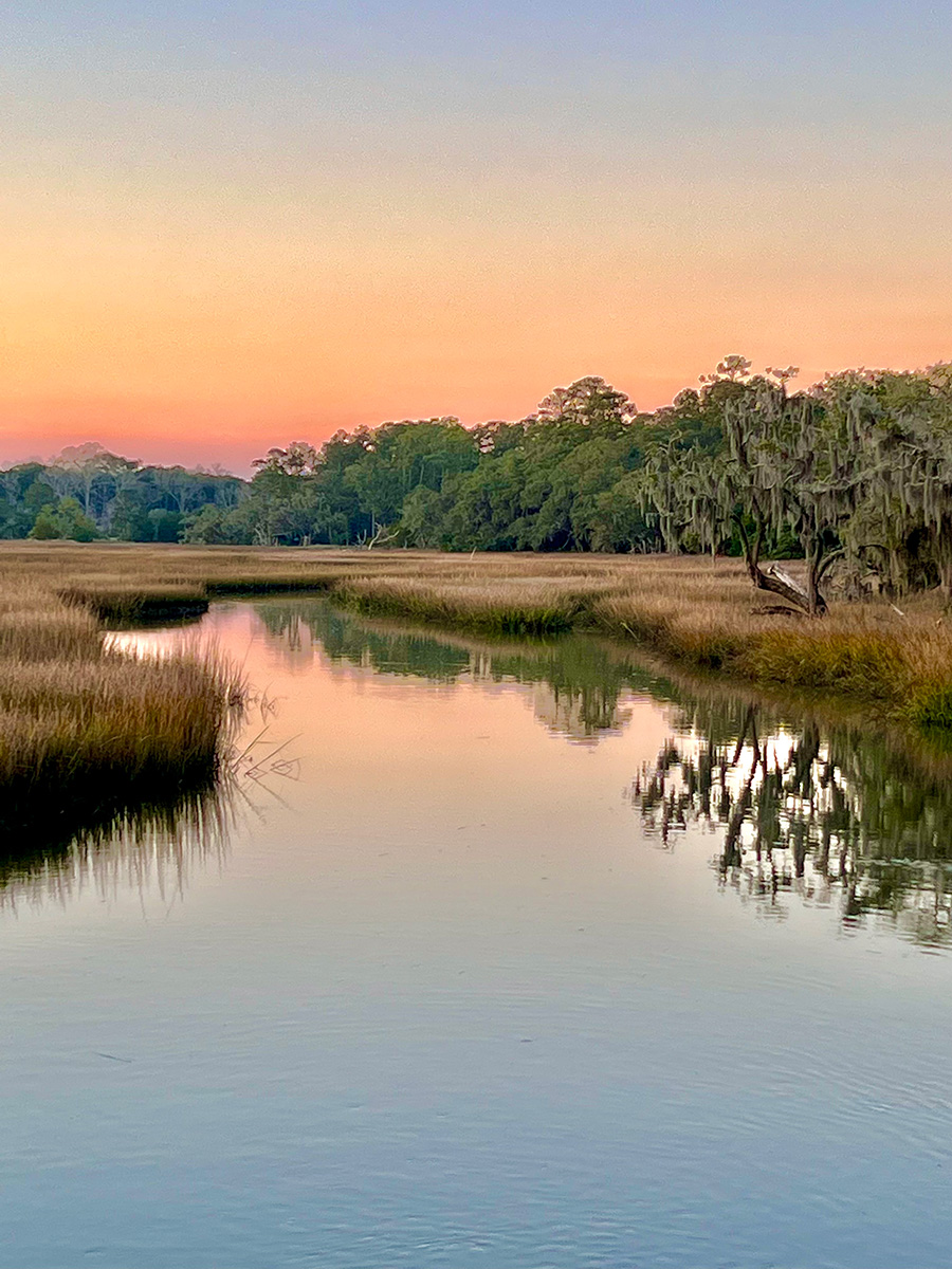 Calm river winding through grassy marshland with trees and a colorful sunset sky.