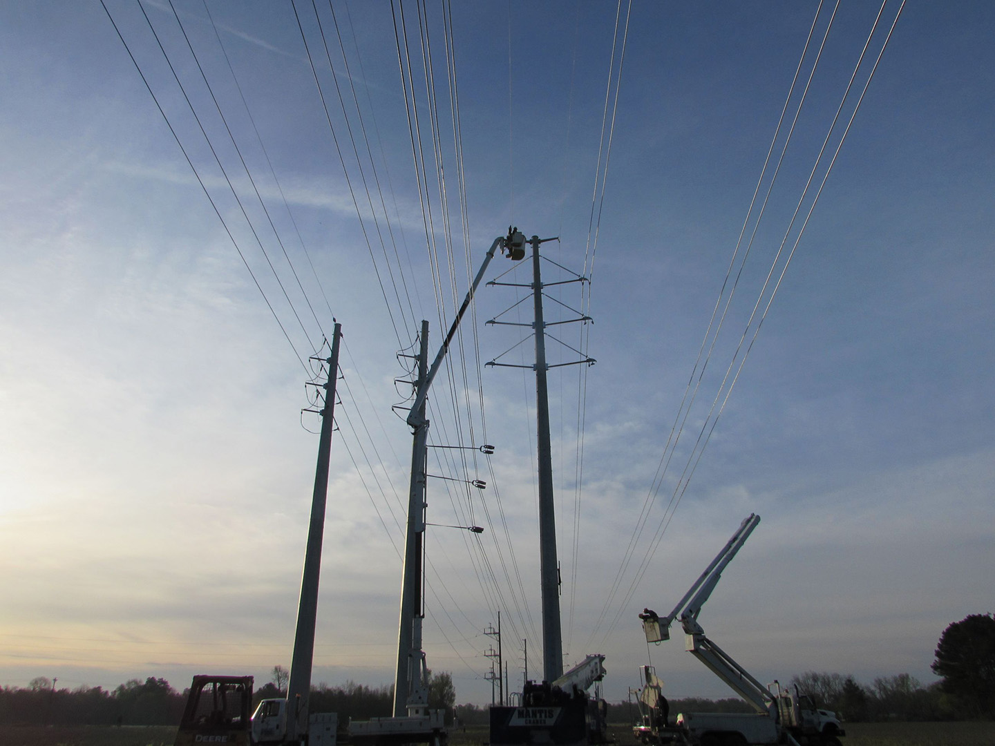 Utility workers in bucket trucks repairing power lines on tall utility poles at dusk.