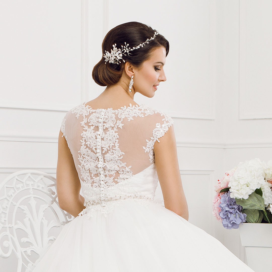 Bride with an elegant hairstyle adorned with a floral hairpiece, wearing a white lace wedding gown with buttons down the back, standing next to a white chair and a vase of colorful flowers.