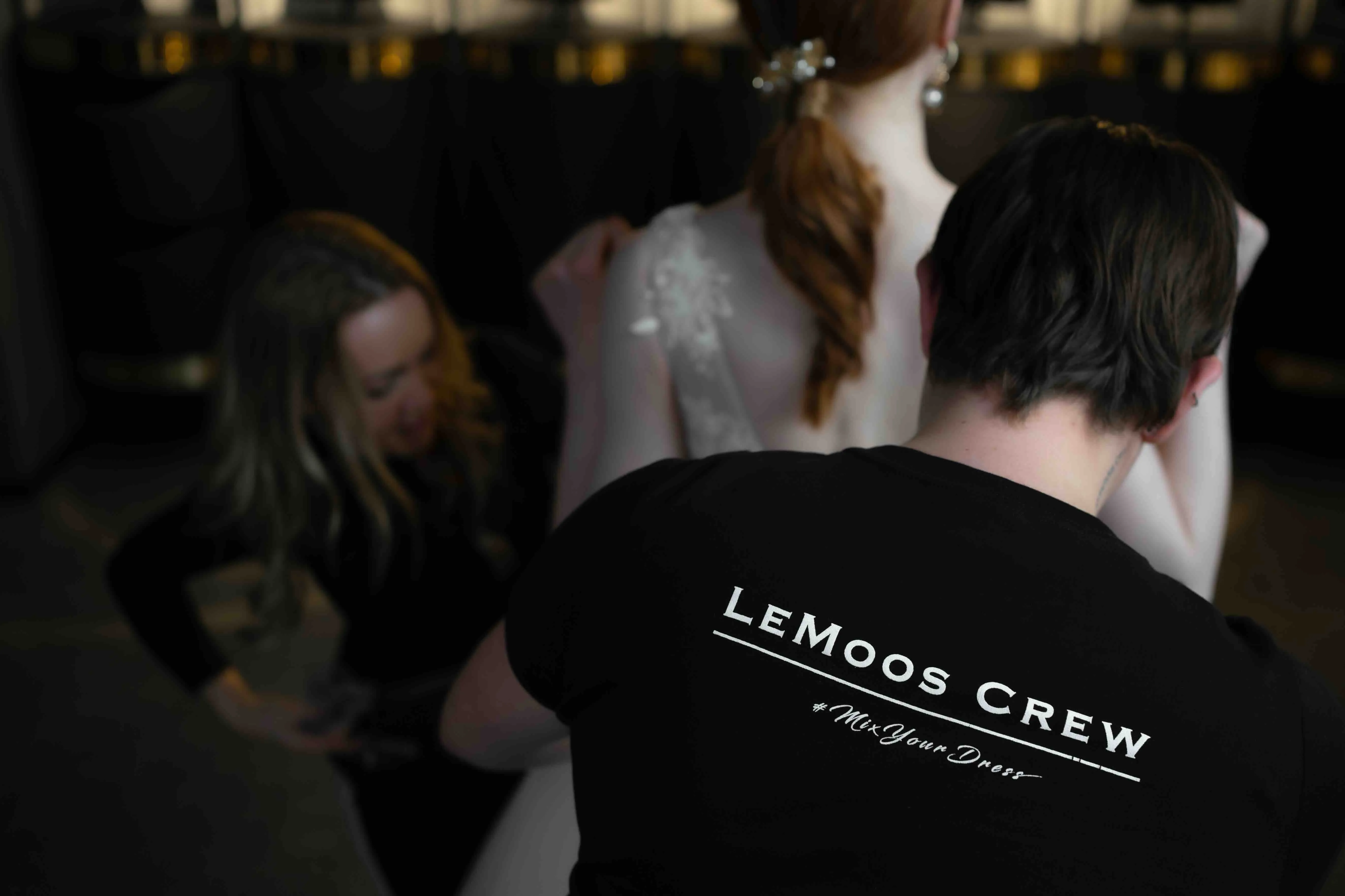 Two people working on fitting a white dress worn by a red-haired woman, one wearing a black shirt with 'LeMoos Crew' printed on the back.
