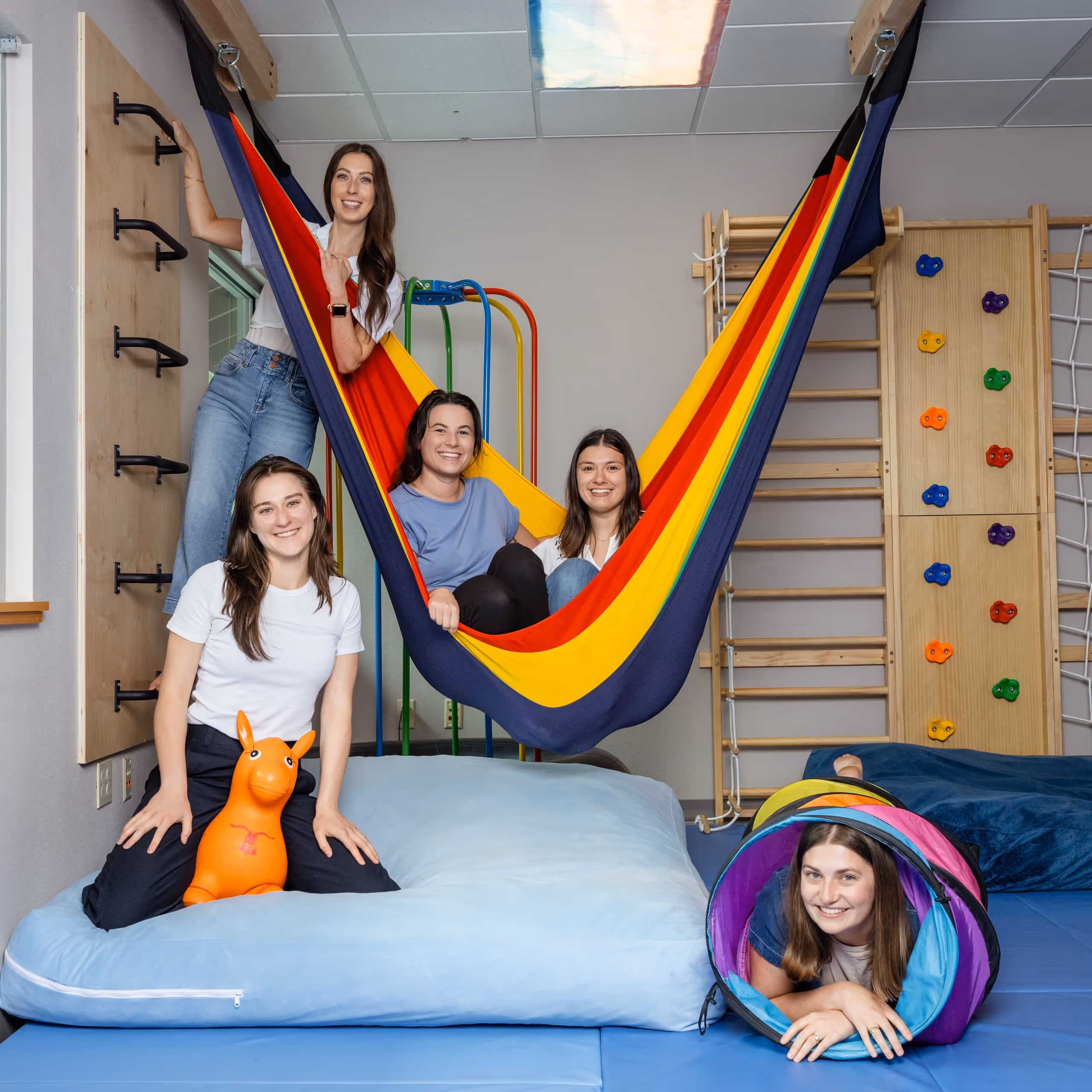 Five women smiling and posing in a colorful indoor playroom with a hammock, climbing wall, tunnel, and soft mats.