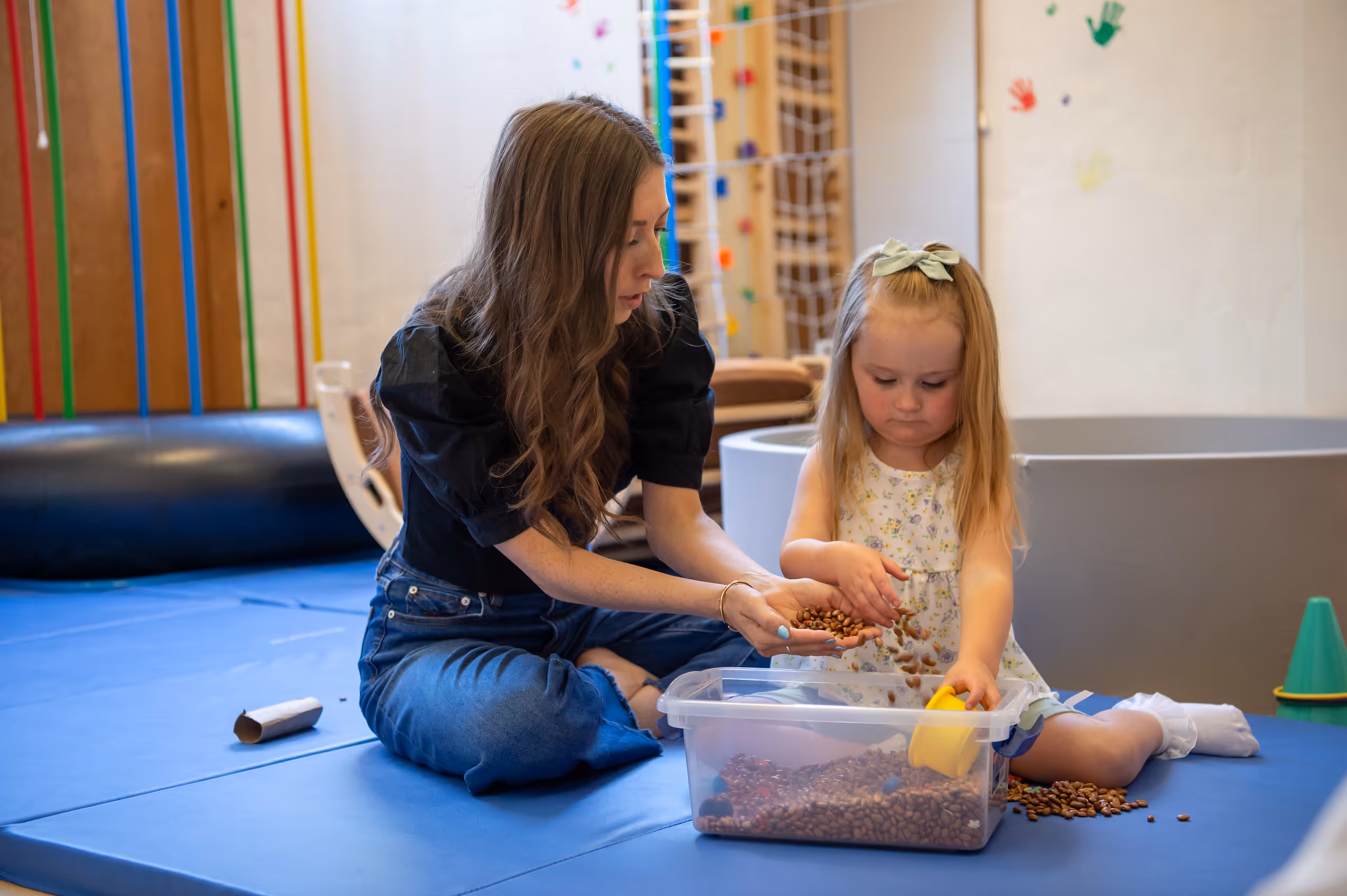 Adult woman and young girl sitting on a blue mat playing with beans in a clear plastic container.