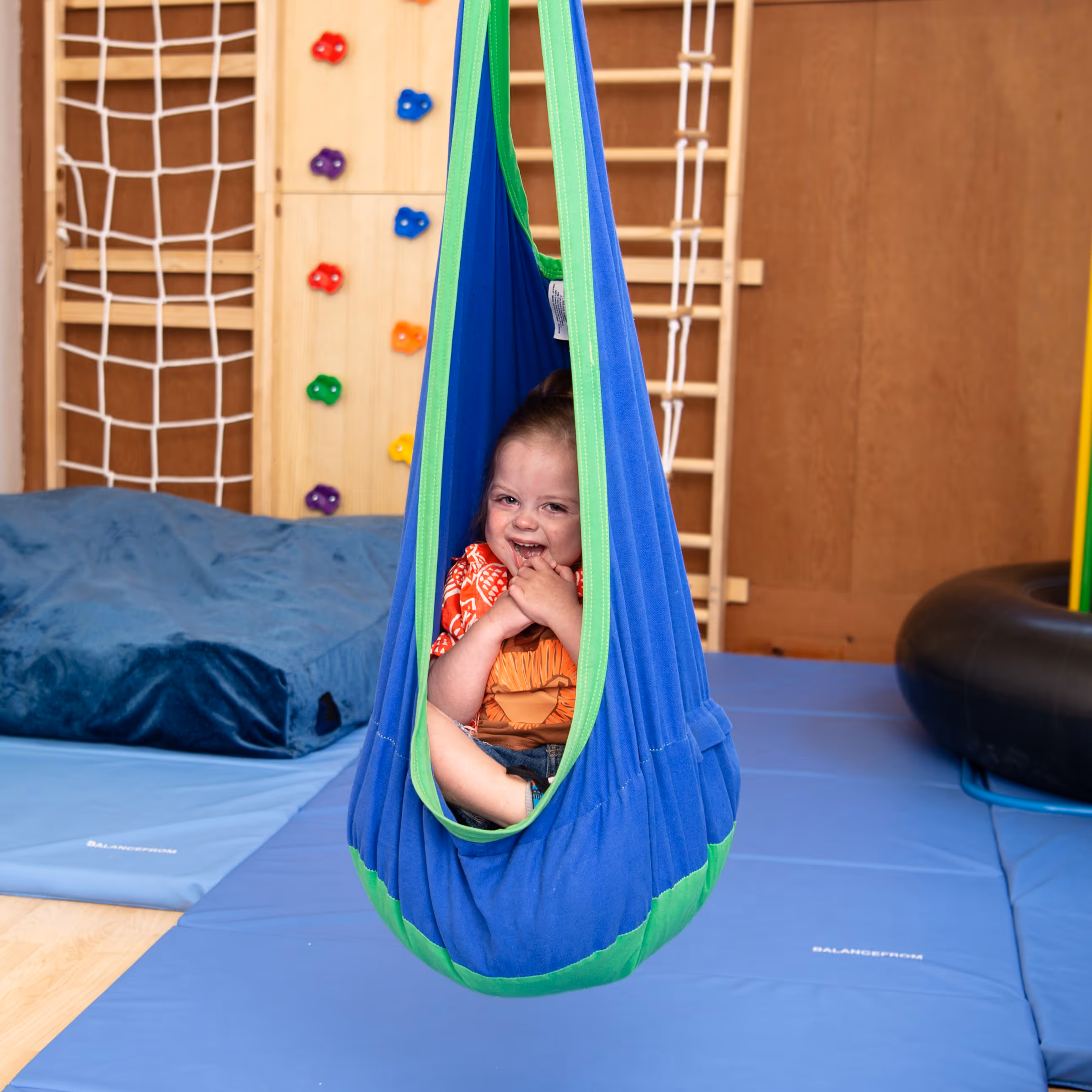 Smiling child sitting in a blue and green sensory swing in a playroom with climbing wall and mats.