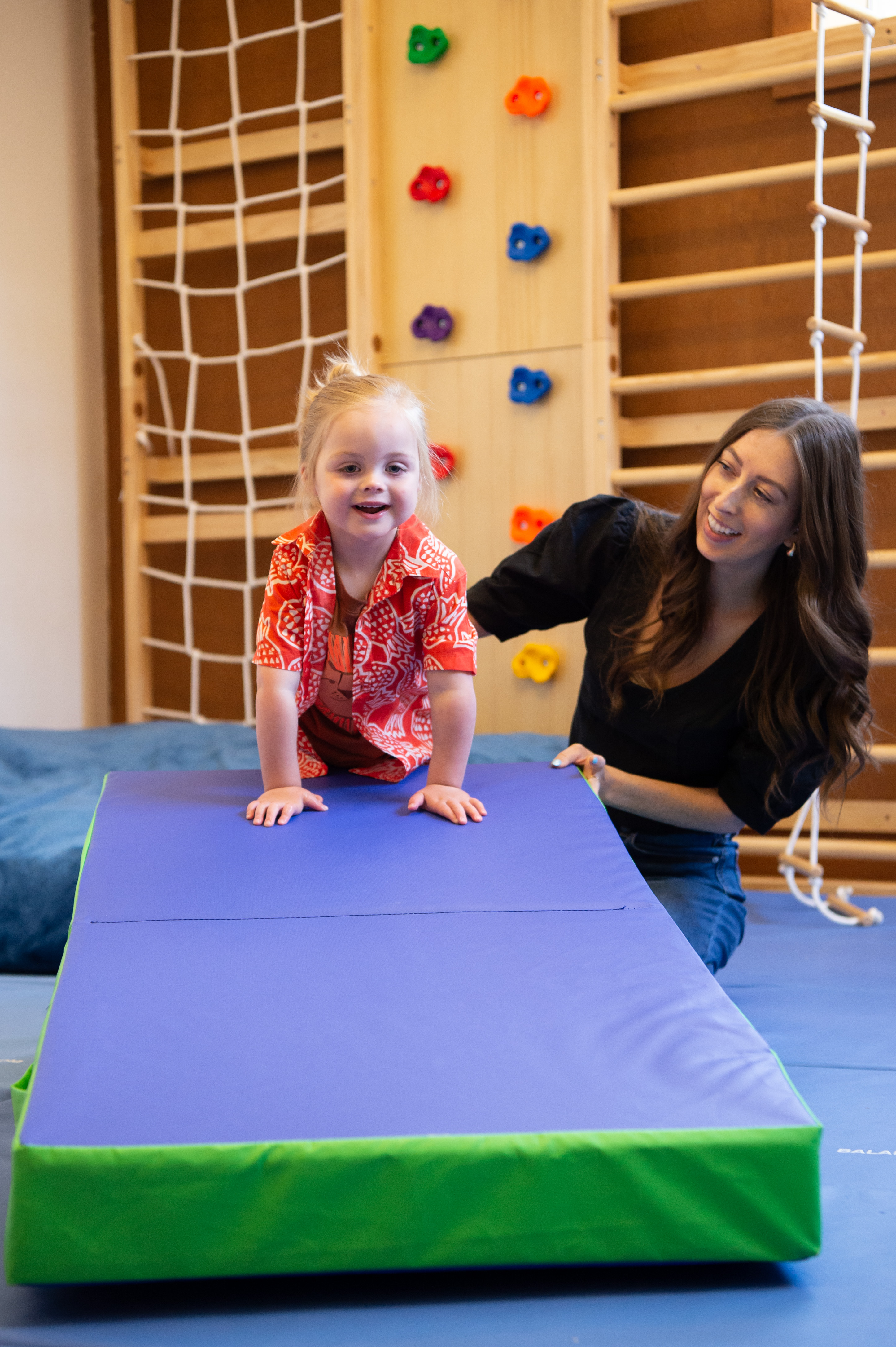 Young girl in red shirt crawling on a purple and green gym mat with a smiling woman supporting her in a playroom with climbing equipment.