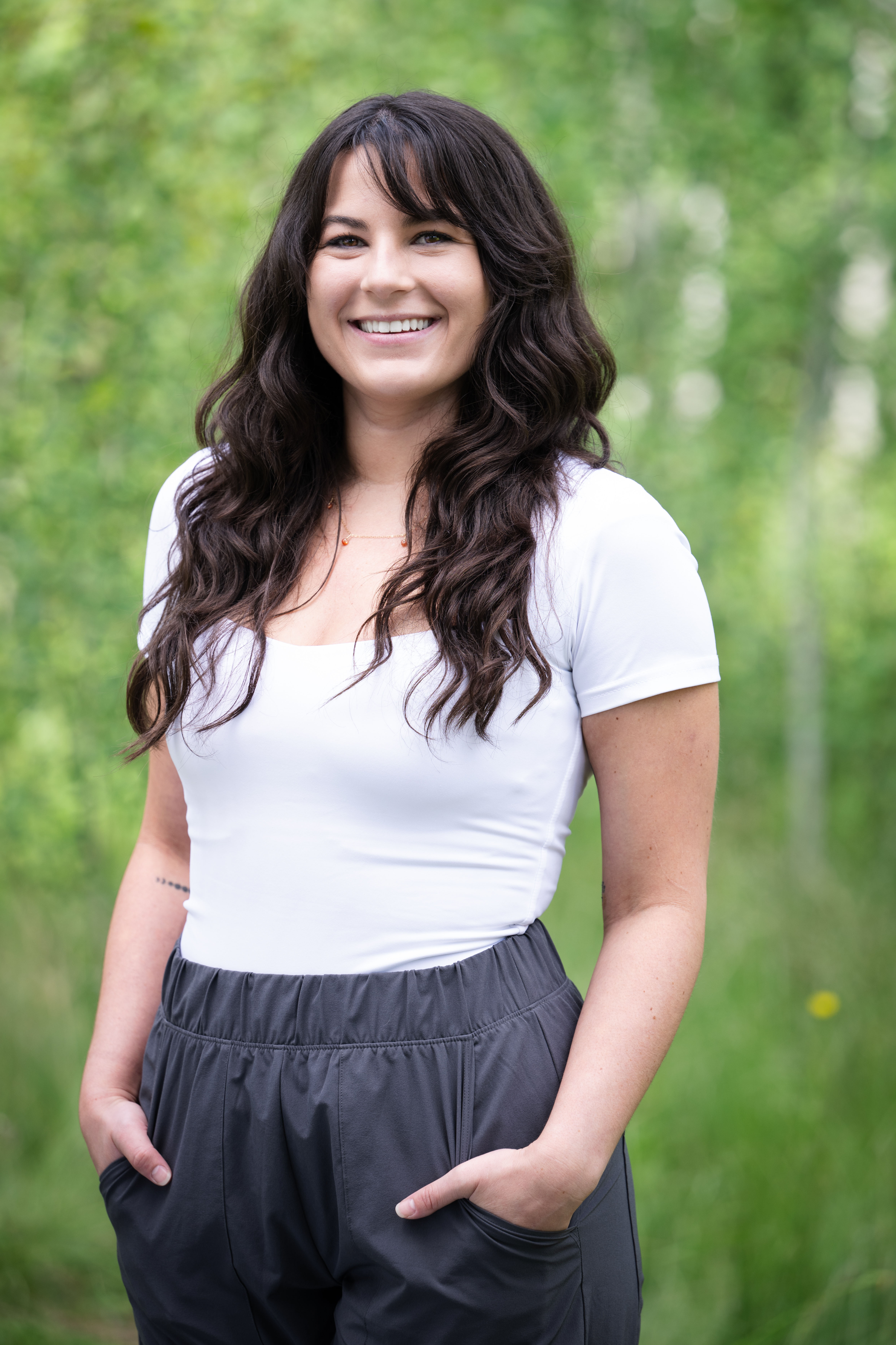 Smiling woman with long wavy dark hair wearing a white top and dark pants, standing with hands in pockets against a blurred green natural background.