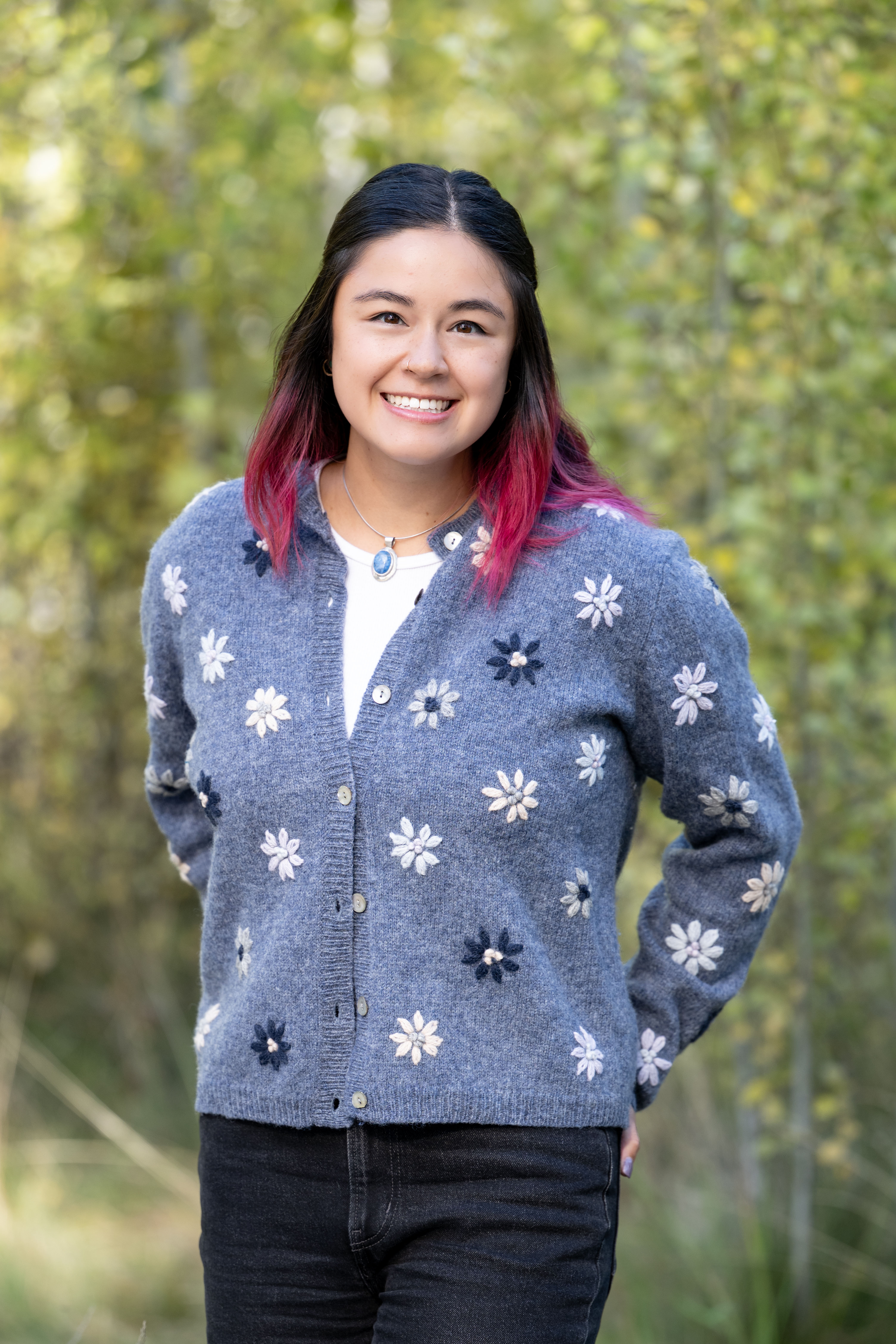 Smiling woman with pink-tipped hair wearing a blue cardigan with white and navy flower patterns, standing outdoors with greenery in the background.