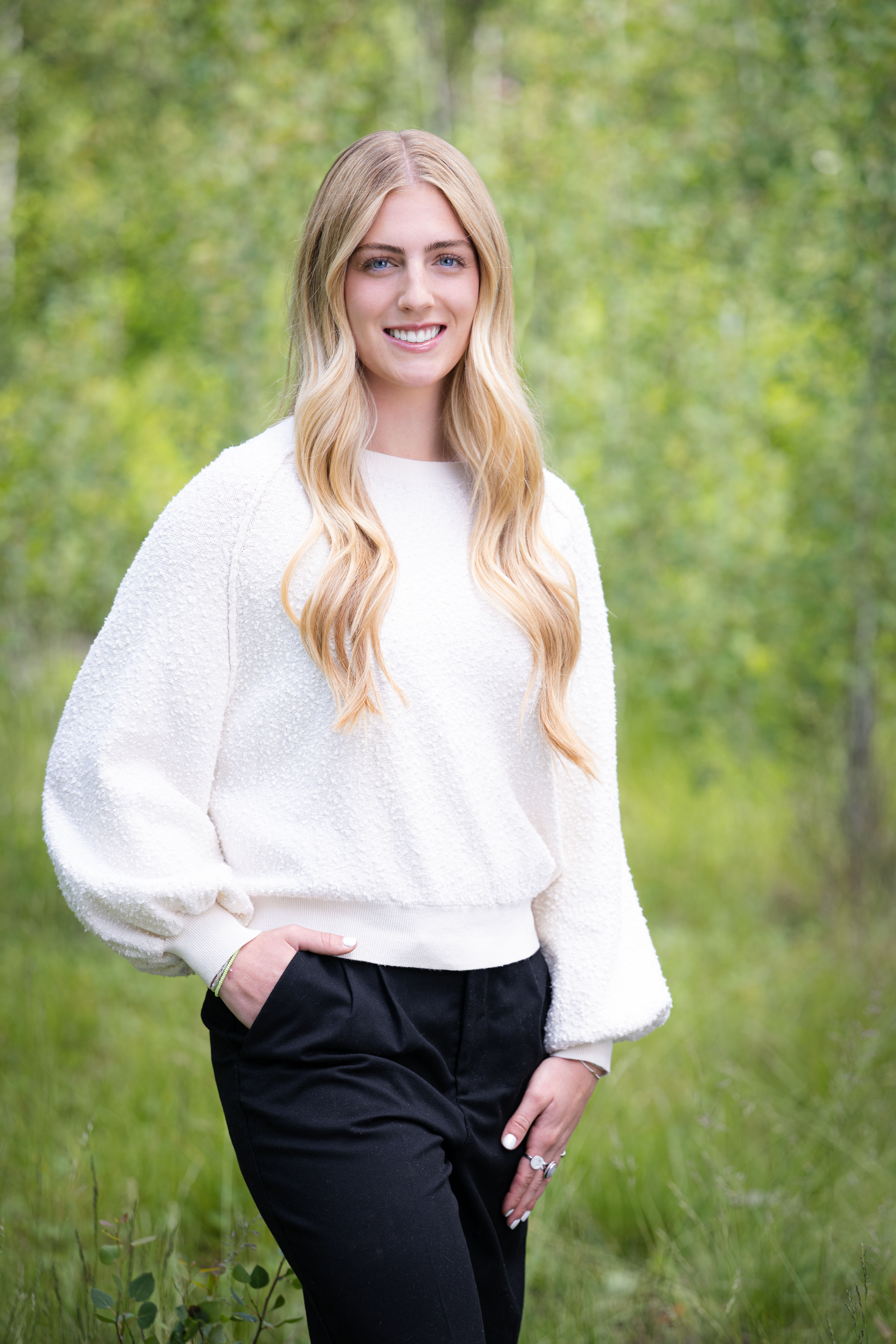 Smiling young woman with long blonde hair in a white sweater and black pants standing outdoors with green foliage background.
