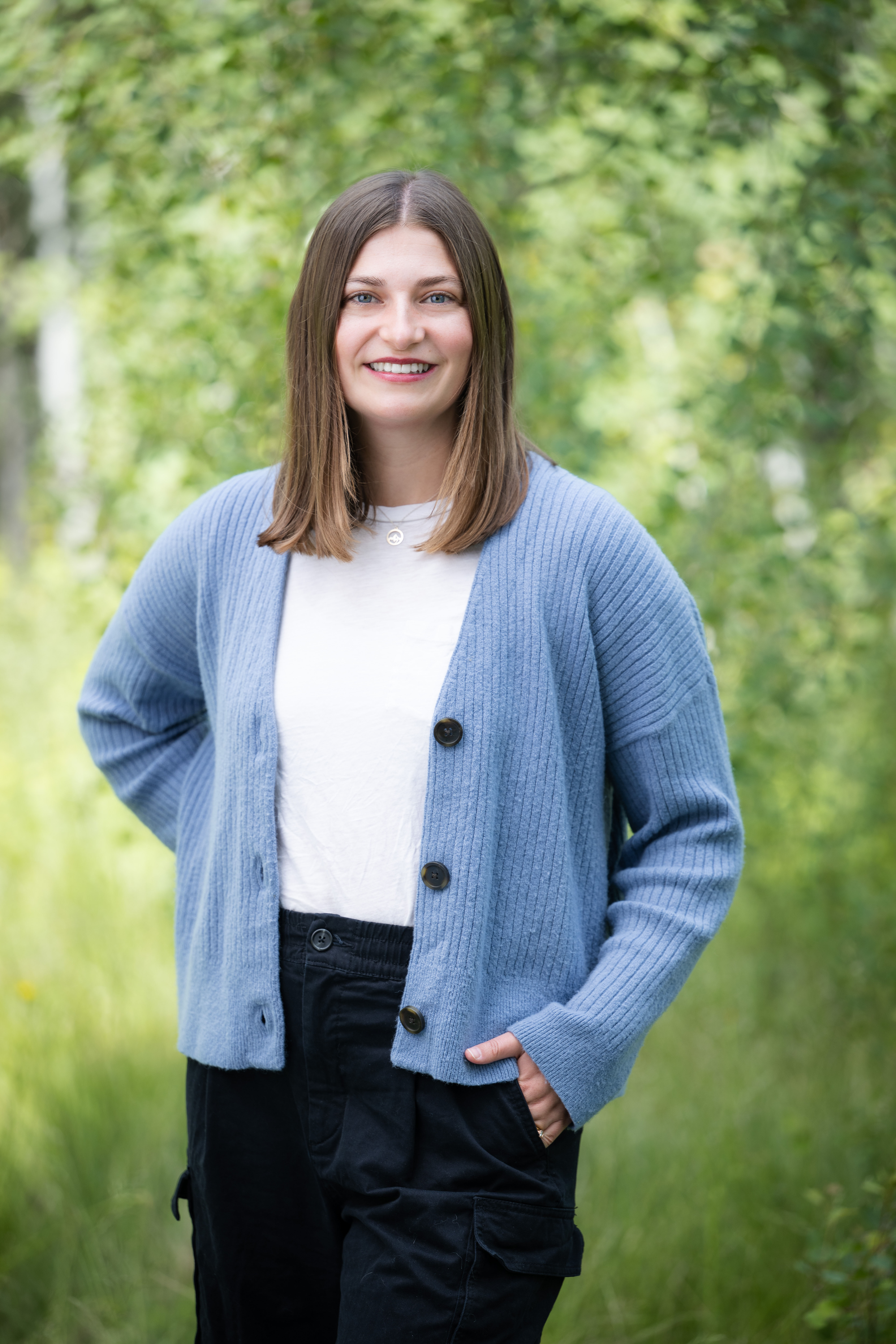 Smiling woman with shoulder-length brown hair wearing a light blue cardigan and black pants standing outdoors with green foliage in the background.