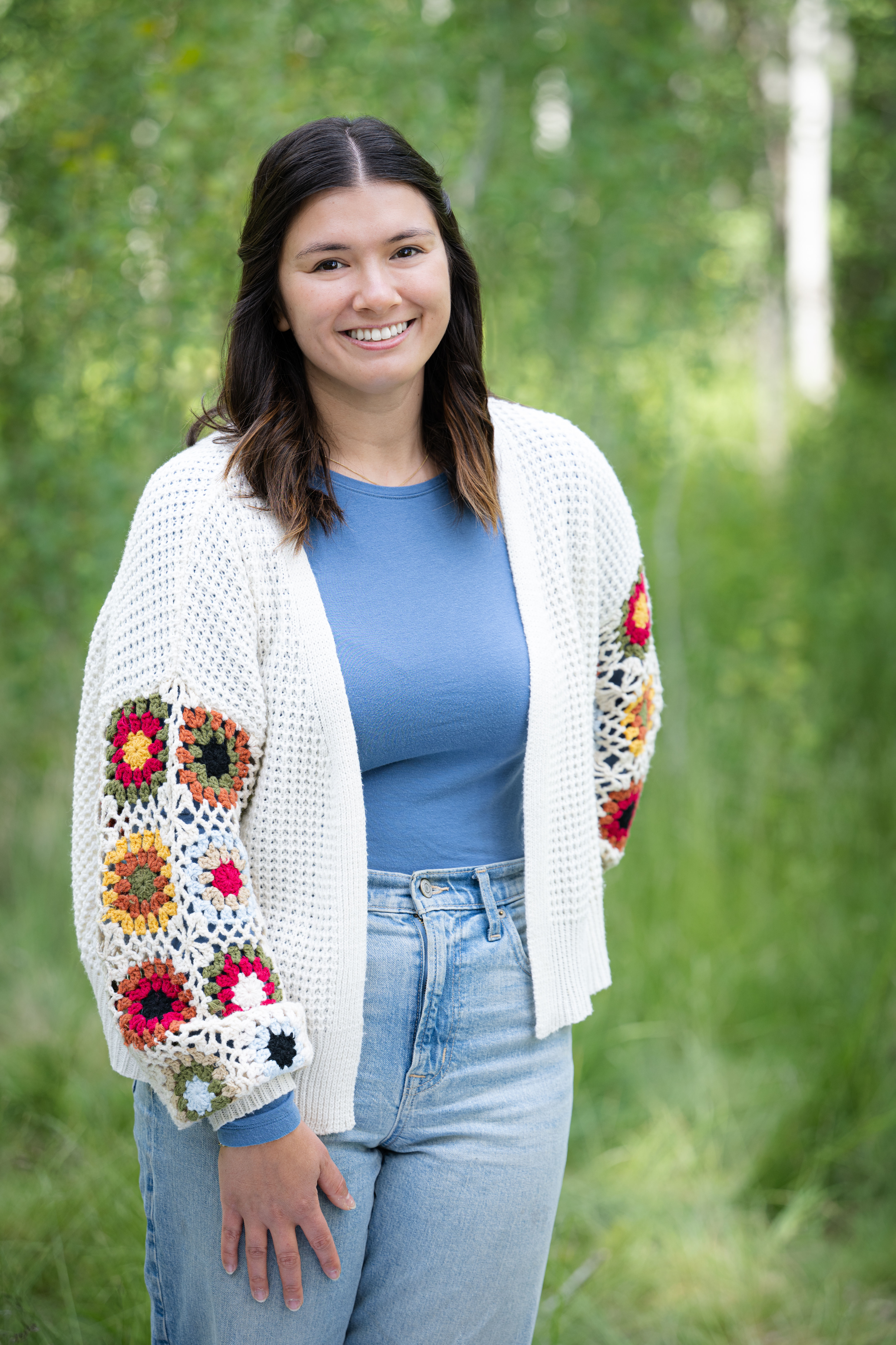Smiling woman with dark hair wearing a blue top, light denim jeans, and a white knitted cardigan with colorful crocheted flower patterns on the sleeves, standing outdoors with greenery in the background.