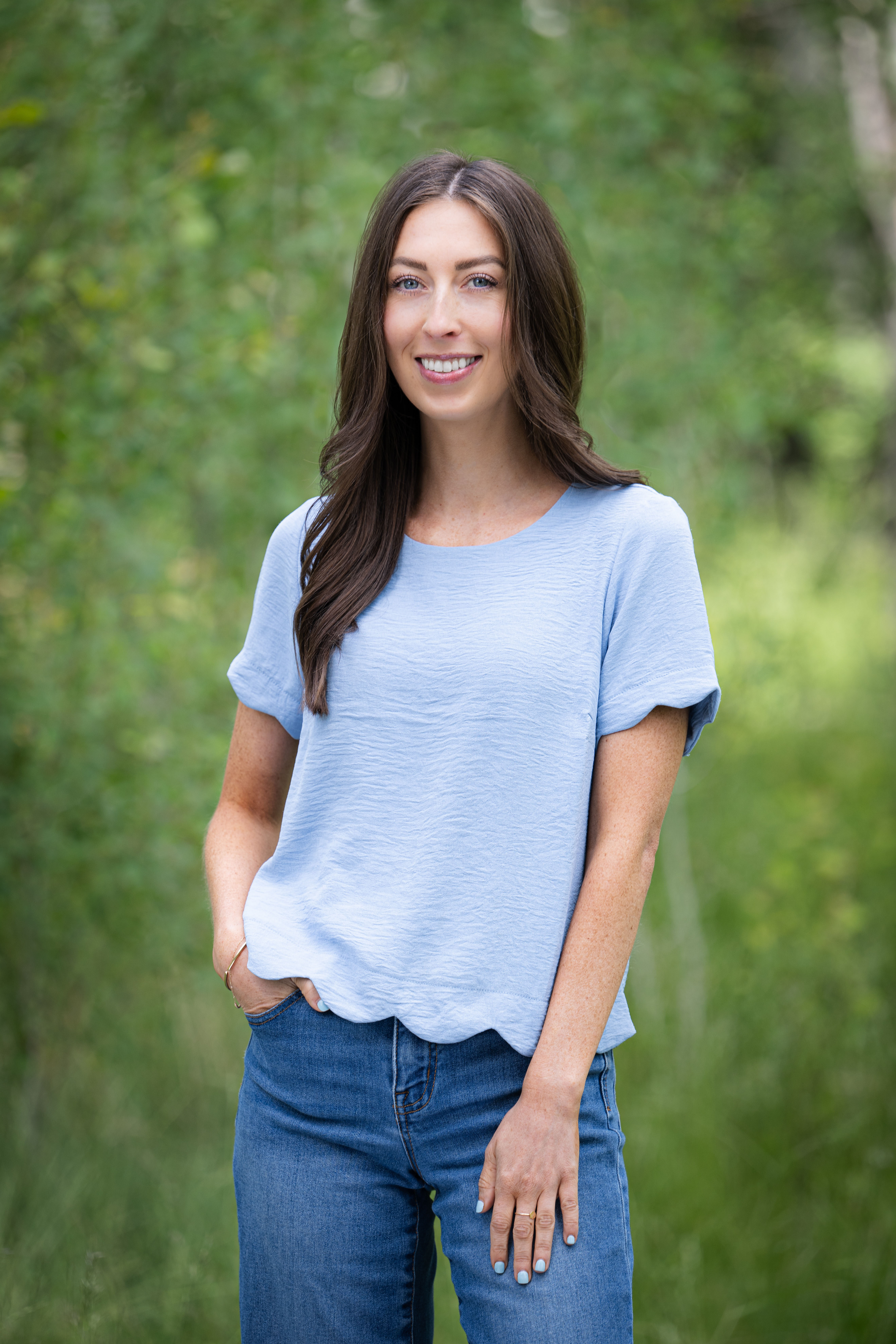 Smiling young woman with long brown hair wearing a light blue shirt and jeans standing outdoors with greenery in the background.