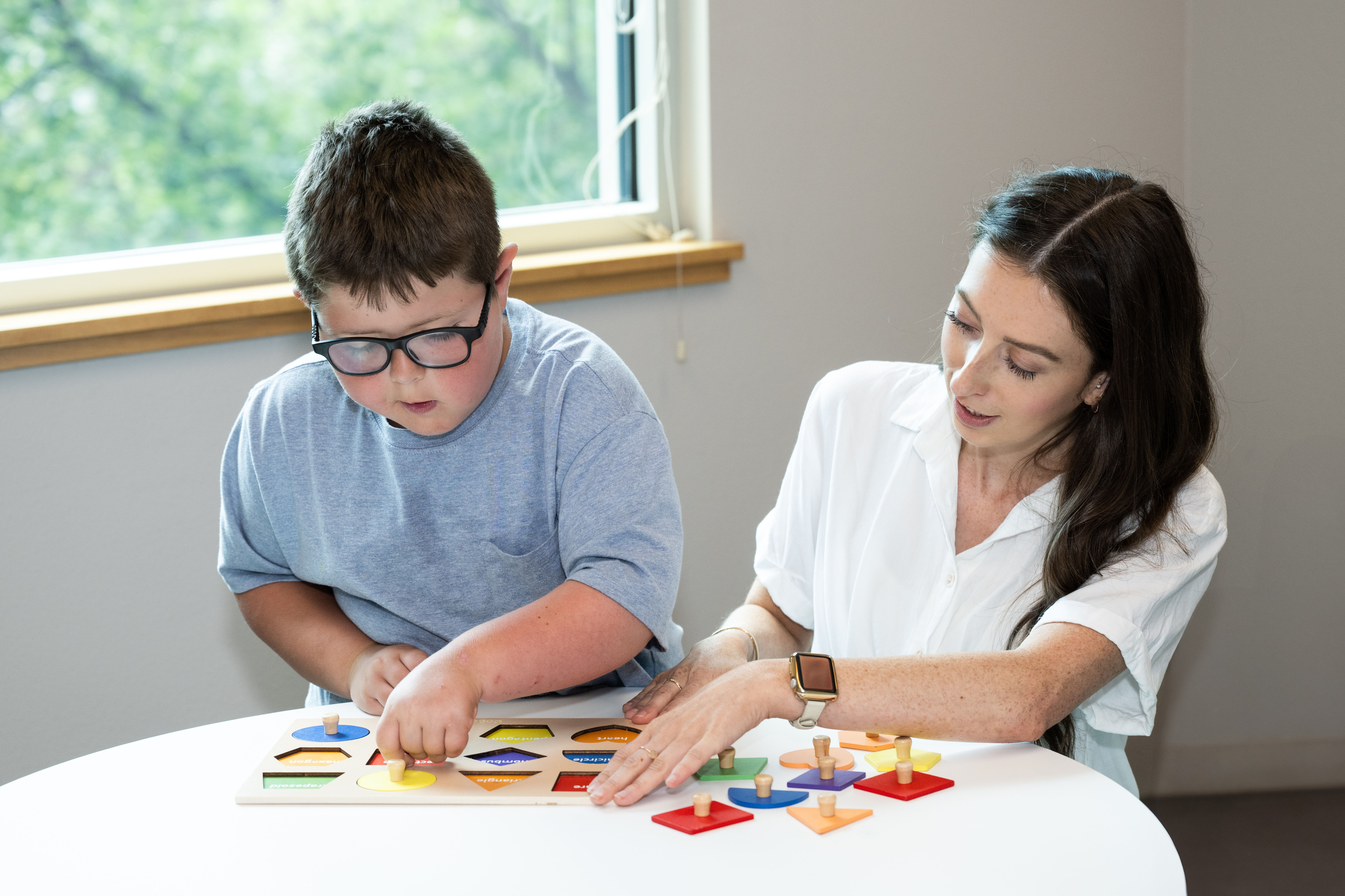 Young boy wearing glasses and a woman playing with a colorful shape puzzle at a white table near a window.