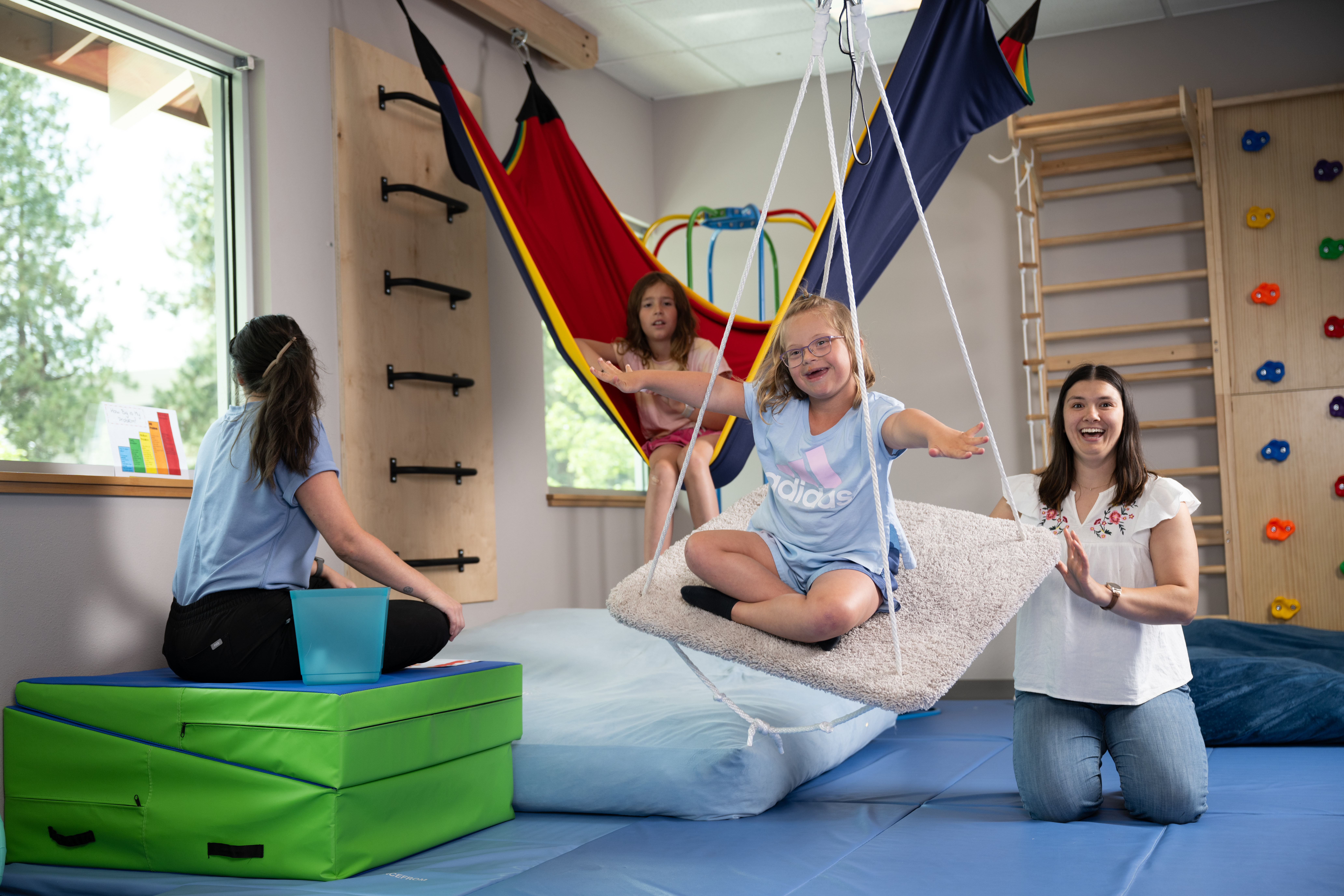 A girl with glasses smiling and sitting cross-legged on a carpeted swing indoors, a woman kneeling nearby supporting the swing, another girl sitting in a colorful hammock, and a person seated on green mats in a playroom with climbing and gym equipment.