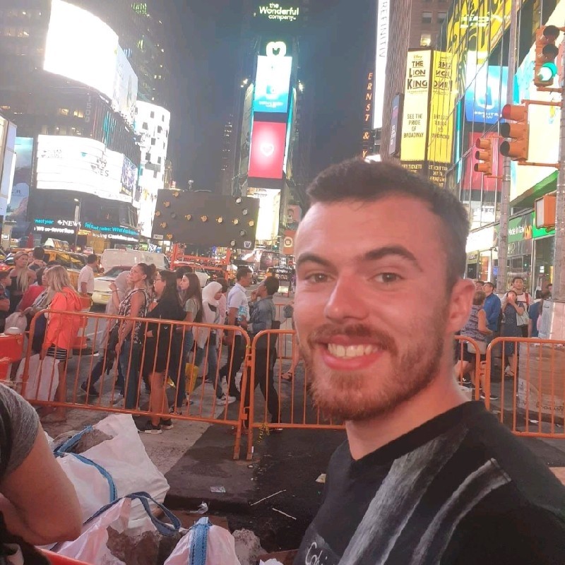 Smiling man with a beard taking a selfie at night in a busy Times Square with crowds and bright billboards.