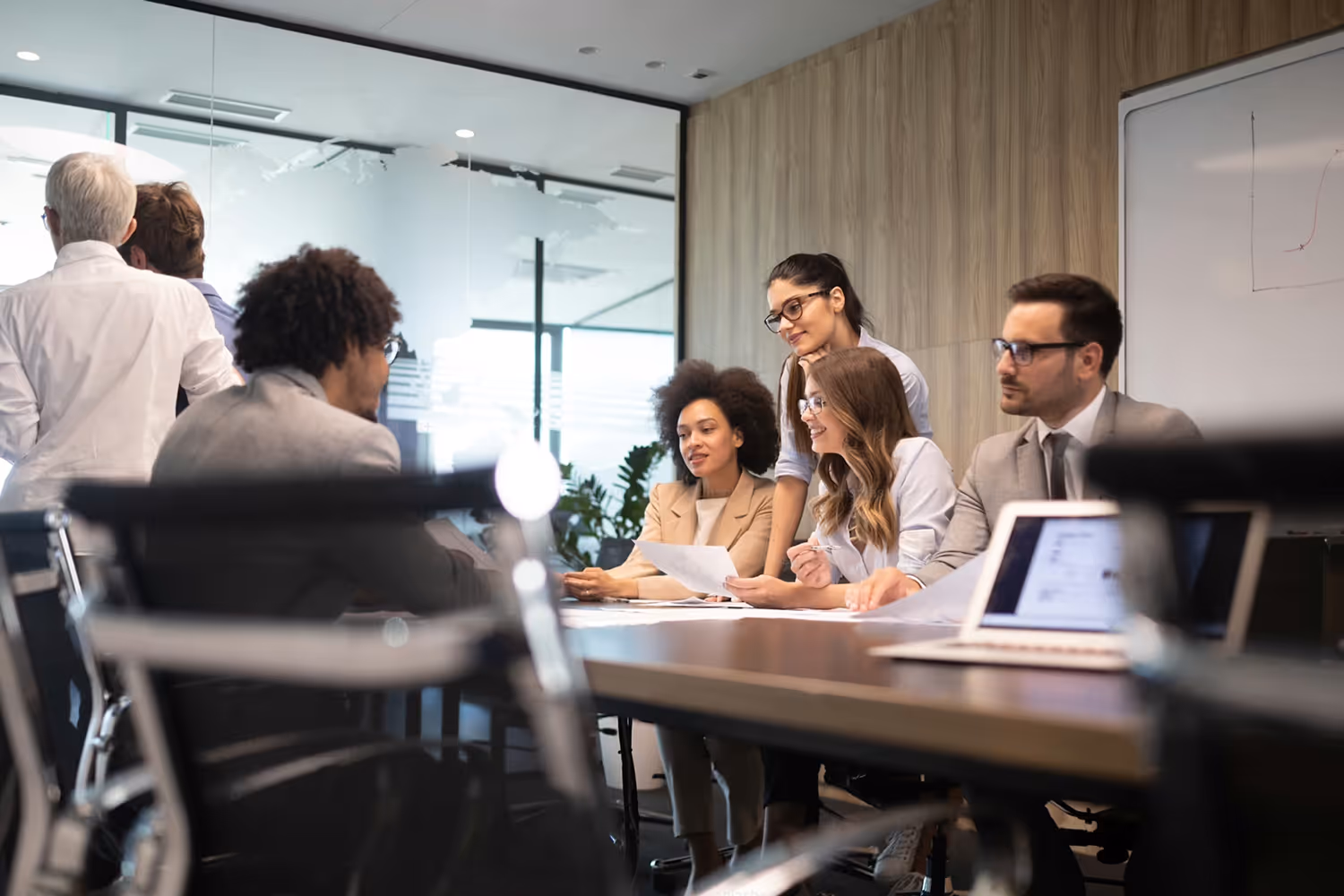 Diverse group of professionals in an office meeting room discussing documents around a table with laptops.