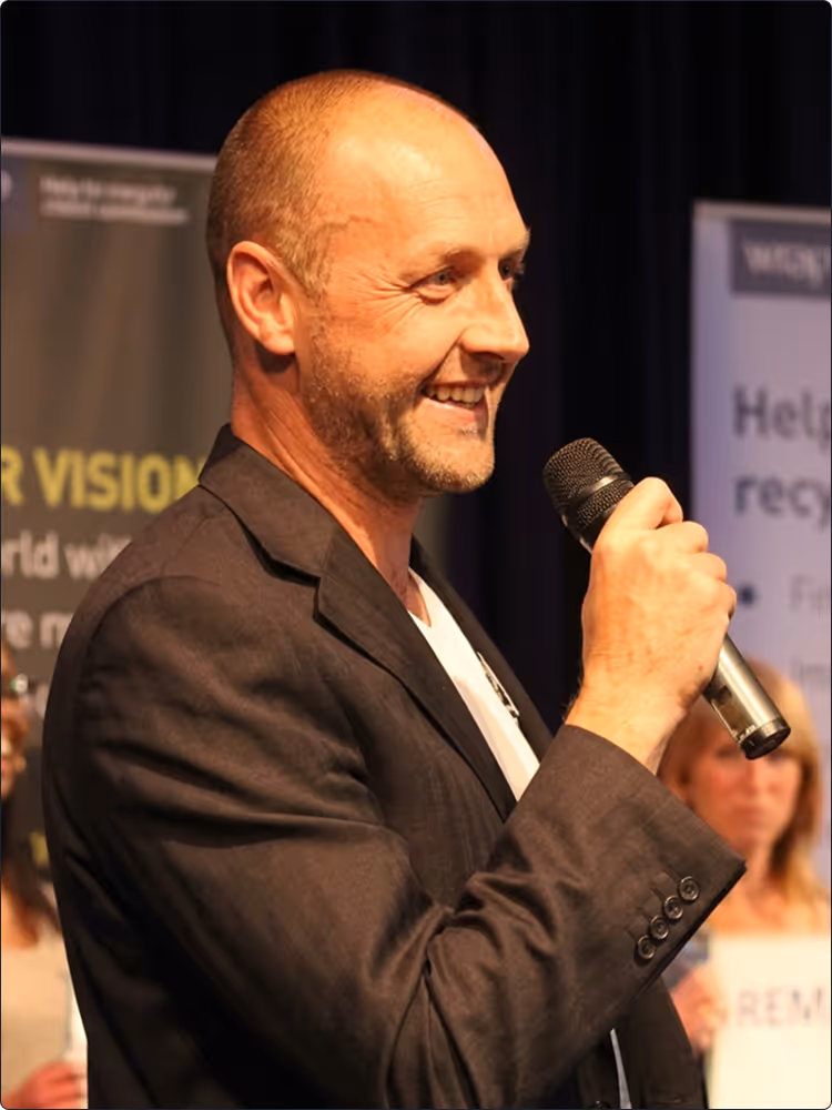 Smiling man in a dark blazer holding a microphone and speaking at an event.