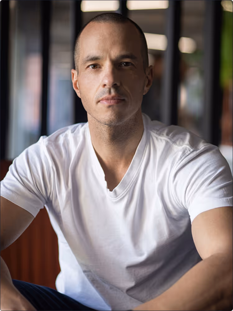 Man with short hair wearing a white t-shirt, looking directly at the camera in an indoor setting.