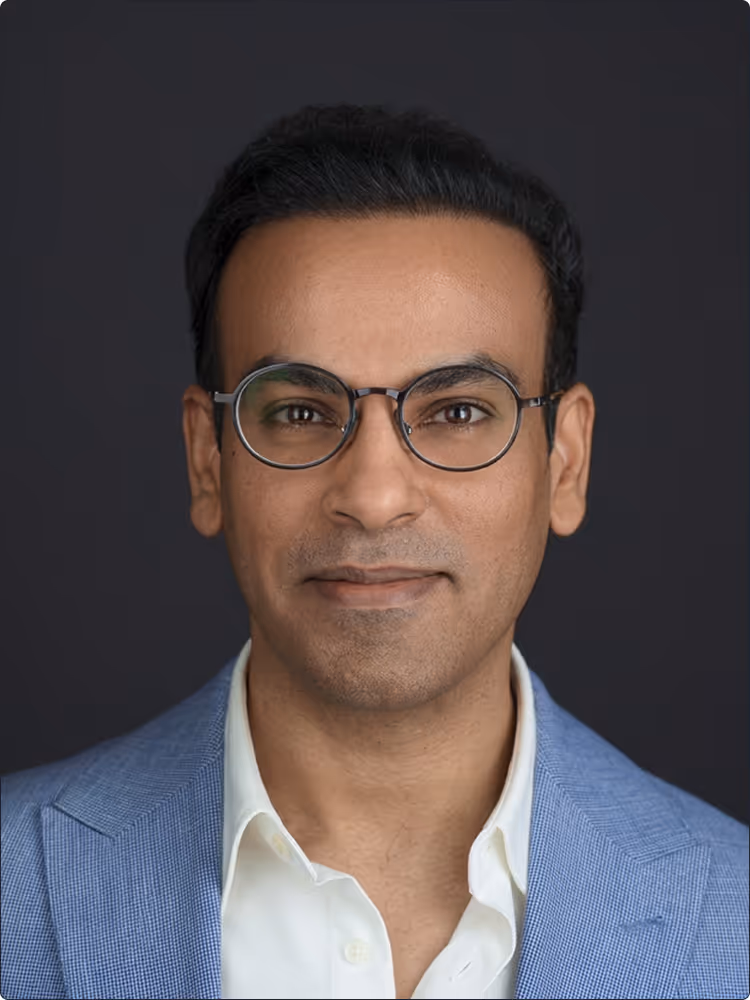 Portrait of a man wearing round glasses, a white shirt, and a light blue blazer against a dark background.