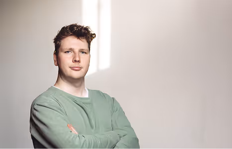 Young man with curly hair in a green sweatshirt standing with crossed arms against a plain light background.