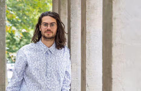 Man with long hair and glasses wearing a patterned shirt, leaning against a light stone pillar with greenery in the background.