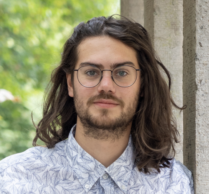 torben Andresen wearing a white shirt with a leaf pattern, standing against a stone wall with green foliage in the background.