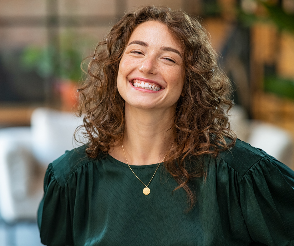 Smiling young woman with curly brown hair wearing a dark green blouse and a gold necklace in a softly blurred indoor setting.