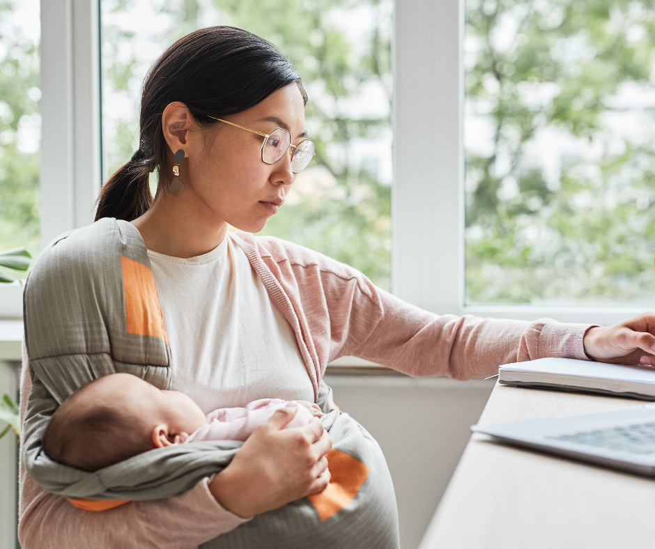 Woman wearing glasses and a pink sweater working on a laptop while holding a sleeping baby in a wrap carrier indoors.