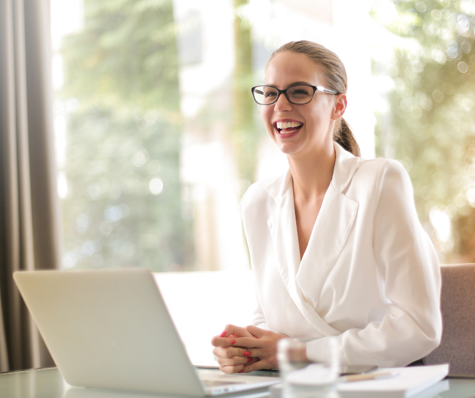 Smiling woman wearing glasses and a white blazer sitting at a desk with a laptop, glass of water, and documents.